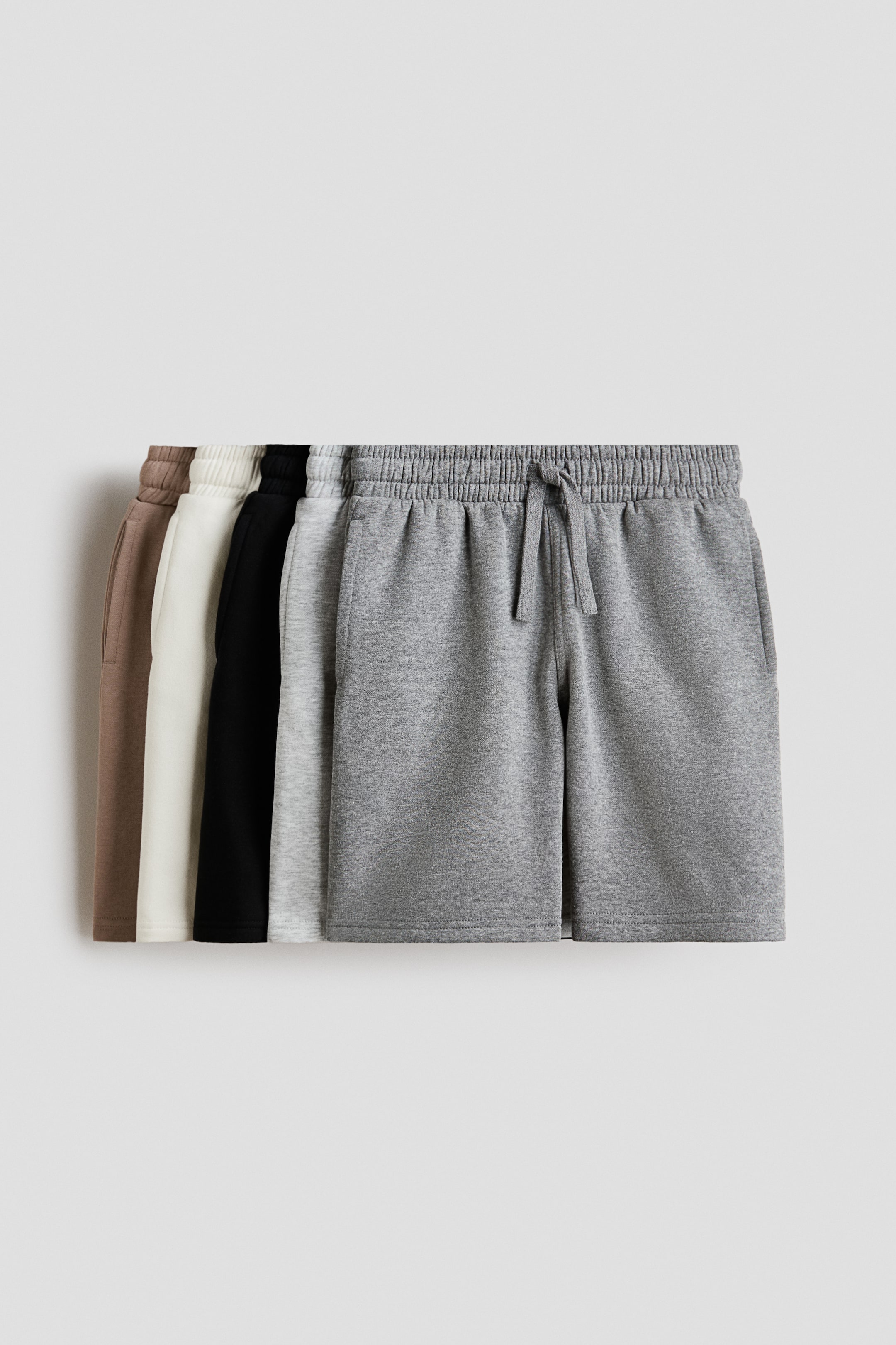 View larger image: "Several pairs of knee-length shorts are showcased from the front, arranged in a variety of colours such as grey, light grey, black, off-white, and brown. Each pair is designed with an elasticated, drawstring waist and subtle side pockets, crafted from a lightweight sweatshirt fabric."