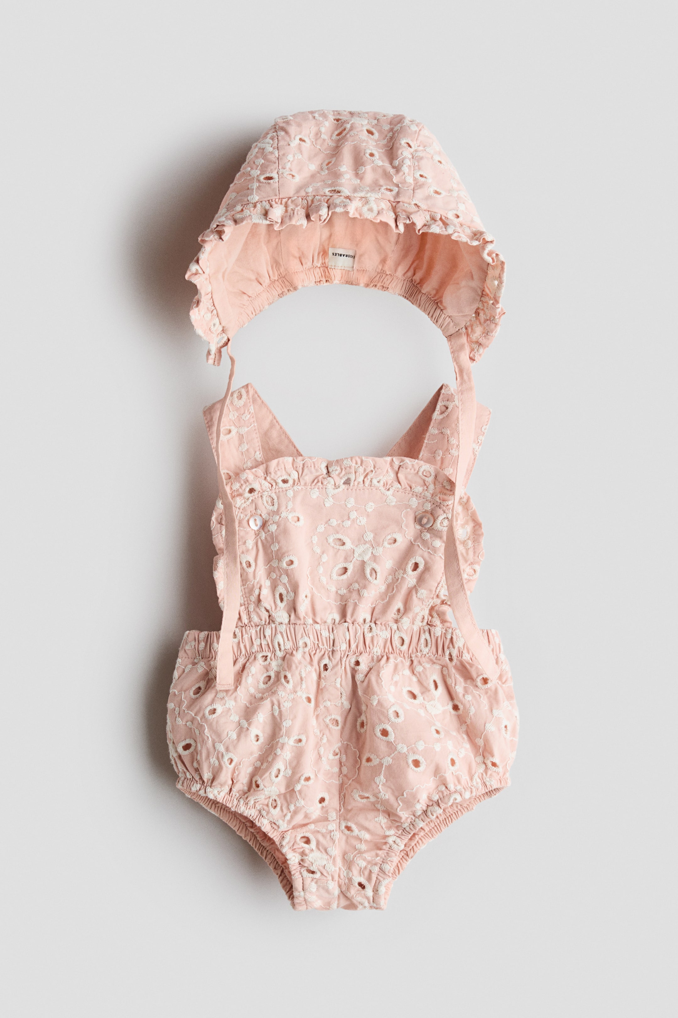 Grösseres Bild ansehen: Ein rosa Lochstickerei-Strampler mit passendem Häubchen-Set wird in der Vorderansicht präsentiert und zeigt weiße Stickmuster und Aussparungen. Der Strampler ist mit einem gerüschten, quadratischen Ausschnitt, Bindeschulterträgern, einem elastischen Bund und elastischen Beinöffnungen gestaltet, während das Häubchen einen gerüschten Rand und Bindebänder aufweist.