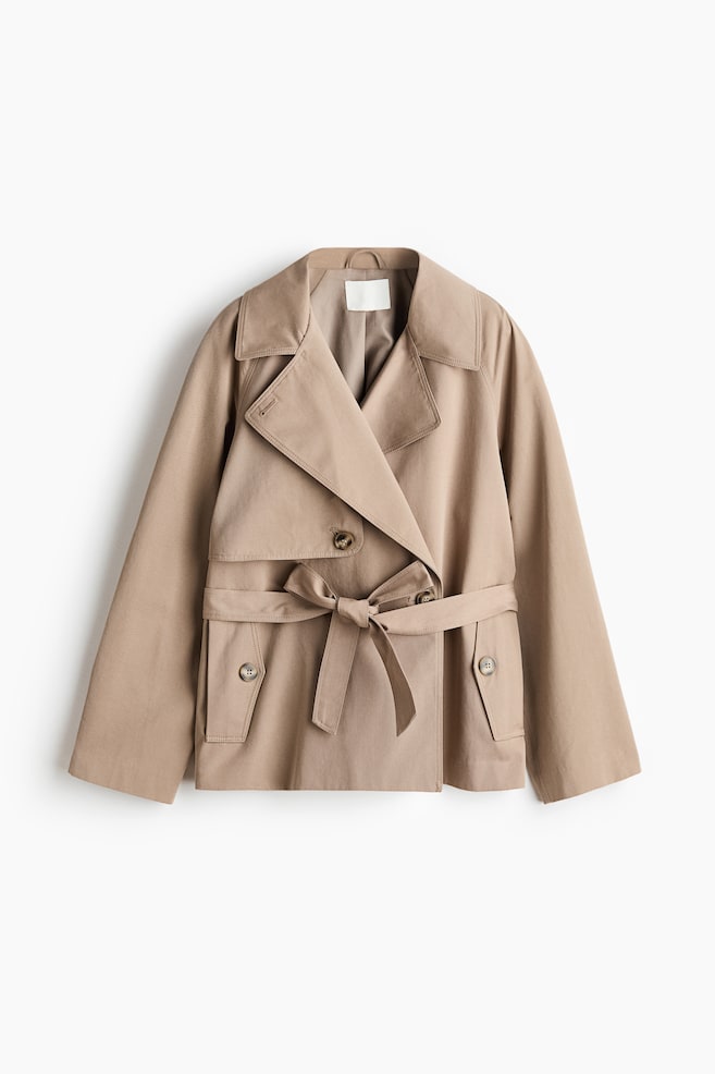 A beige cropped trench coat is displayed flat from the front, featuring a double-breasted design with wide lapels, an adjustable belt tied in a bow at the waist, and front flap pockets with buttons. - 1