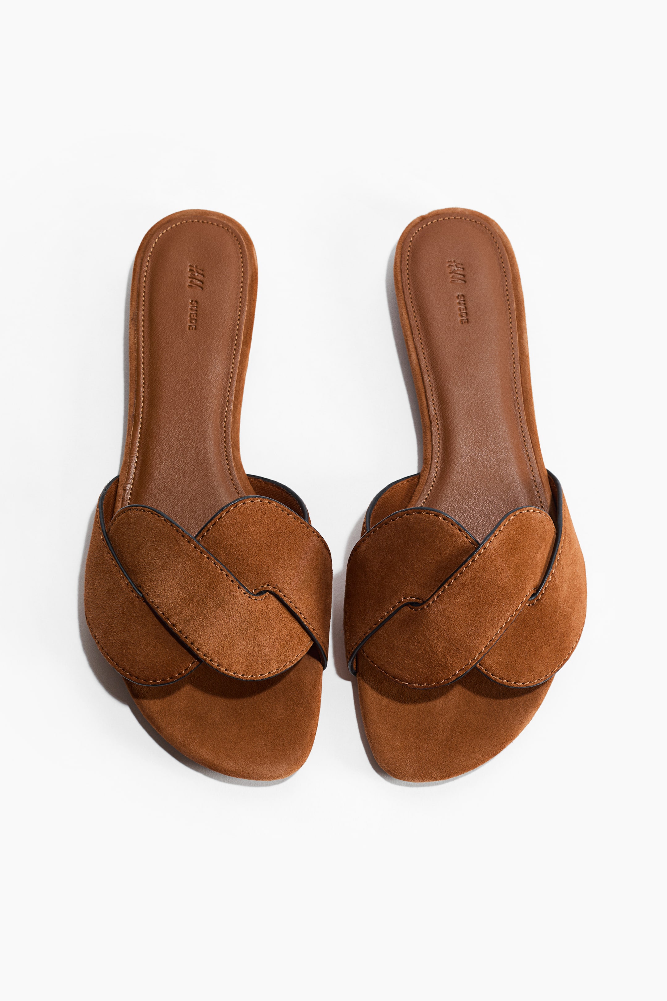 View larger image: Brown suede slip-on sandals are showcased FRONT, featuring wide, overlapping straps with twisted detailing across the vamp, outlined with dark piping. The flat brown footbeds display visible stitching and "H&M SUEDE" debossed near the heel.