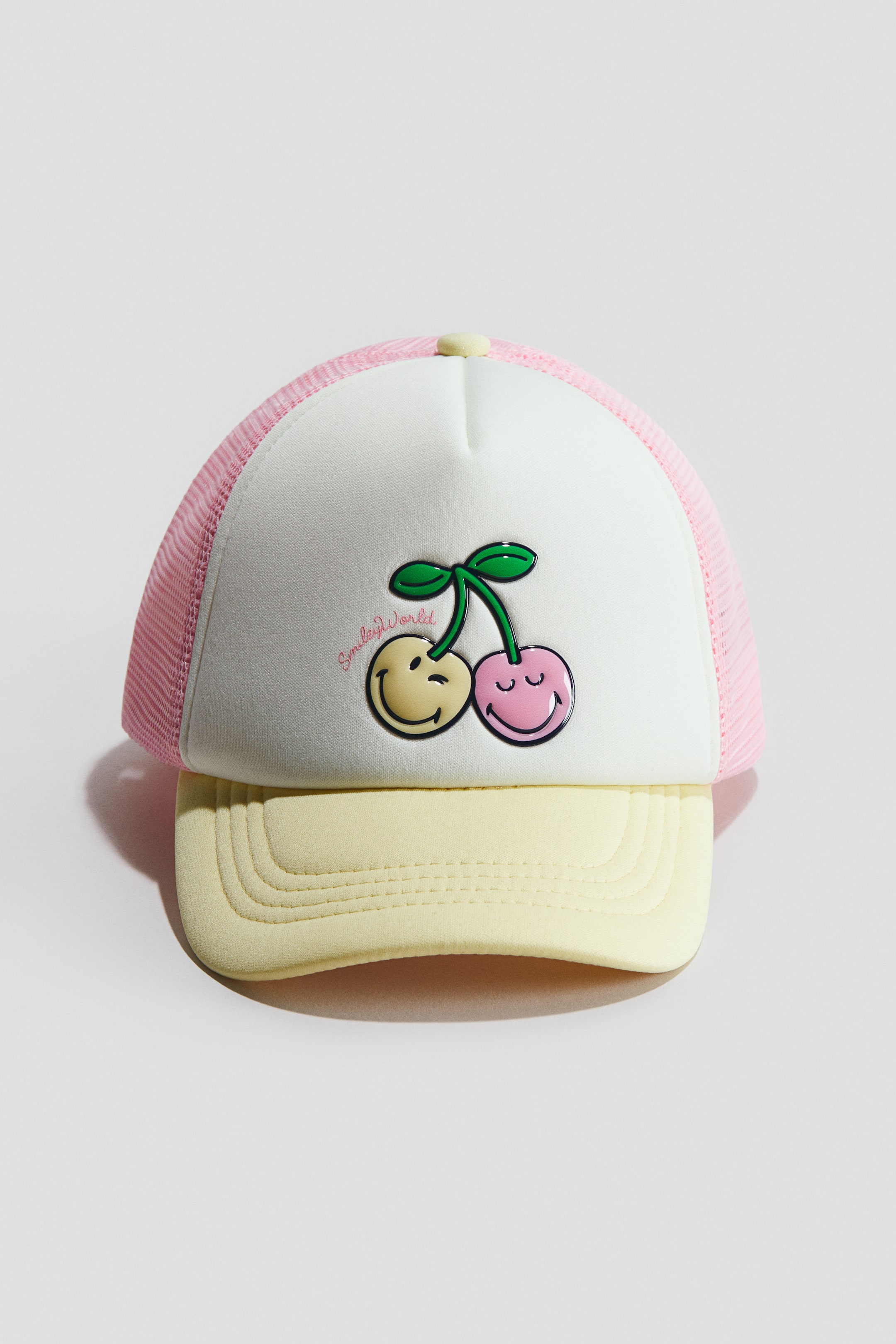 View larger image: A trucker cap is showcasing FRONT, featuring a white woven front panel adorned with a printed design of two cherries; one yellow with a winking smiley face and one pink with a smiling face, connected by green stems with 'SmileyWorld' printed below. Its pink mesh back and curved yellow brim with stitched detailing are also visible.