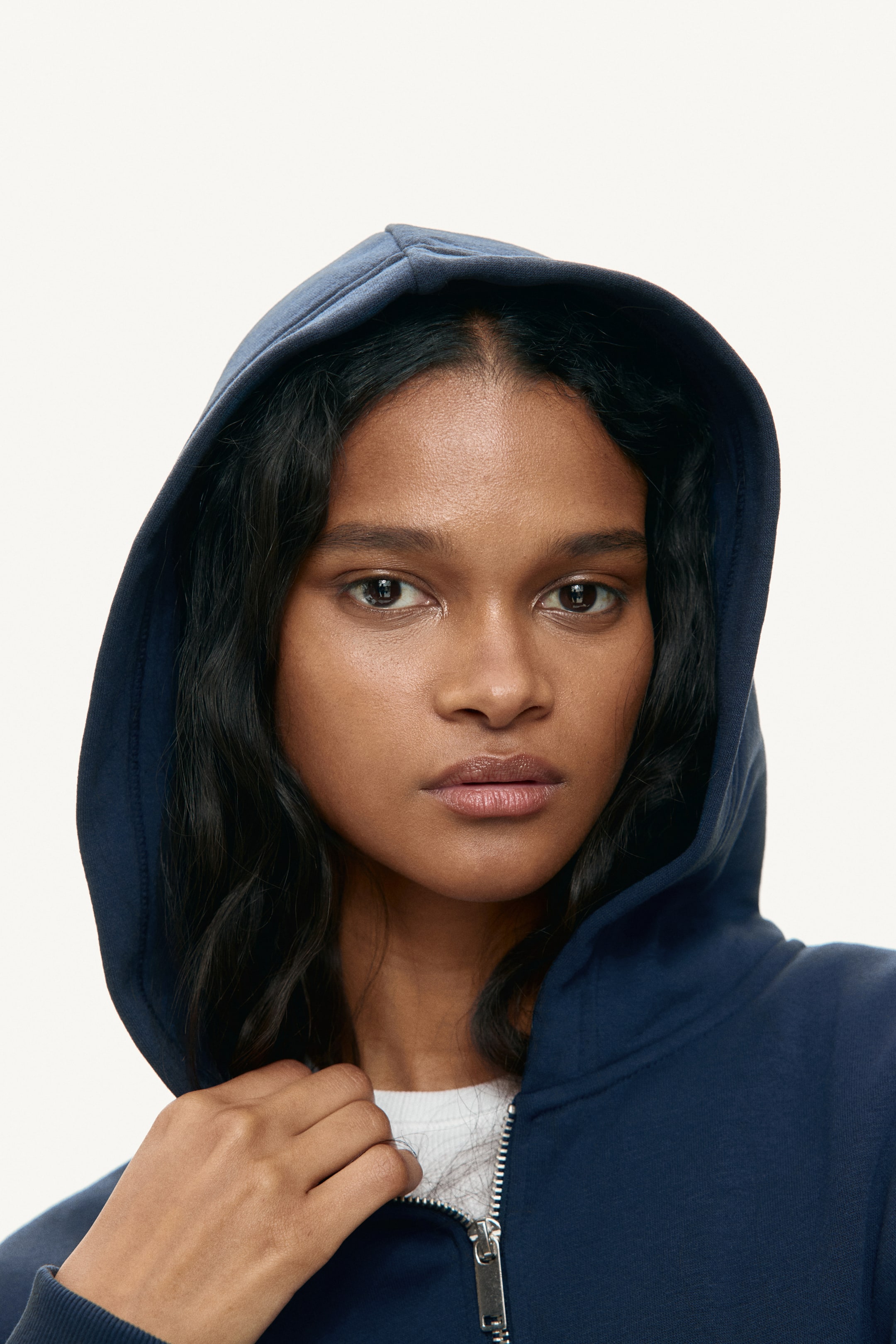 Voir une image plus grande: La mannequin porte un chandail à capuche bleu marine avec fermeture éclair à l'avant, sa capuche couvrant sa tête. Elle tient le devant du chandail, près de la fermeture éclair partiellement ouverte et du poignet côtelé, révélant un t-shirt blanc en dessous.