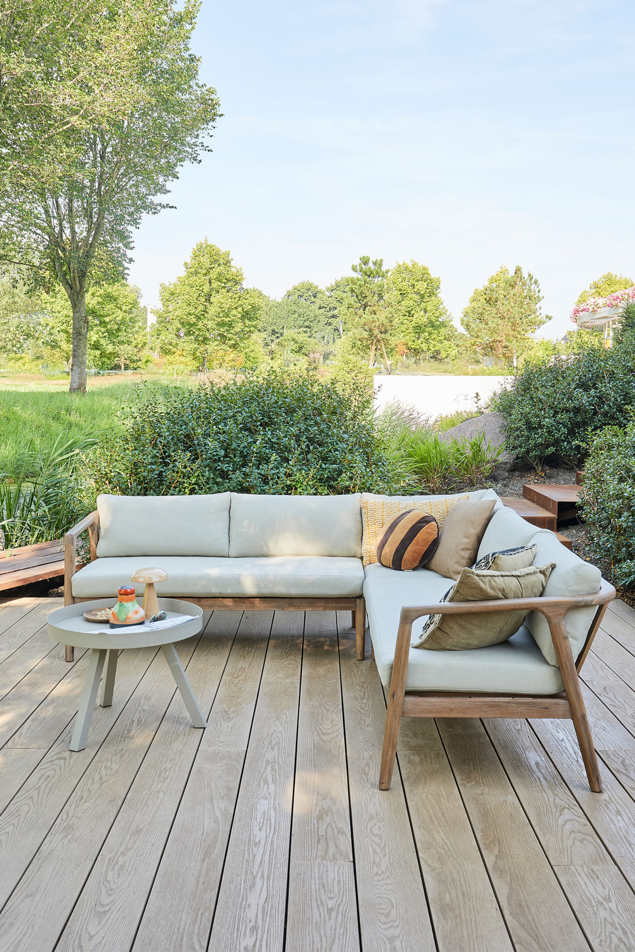 Agrandir l'image: Un canapé d'angle extérieur en forme de L, doté d'un cadre en bois clair et de coussins beige clair, est disposé avec sa section la plus longue s'étendant vers la gauche et sa section de retour plus courte vers la droite. Plusieurs coussins décoratifs reposent sur les coussins, et une petite table d'appoint ronde gris clair à trois pieds est placée à côté du canapé, présentant un objet en forme de champignon, une cruche colorée et un petit plat.