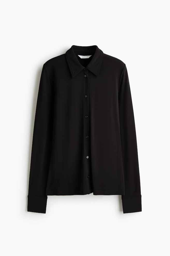 A black fitted shirt is hanging front-facing, showcasing a pointed collar, a central button fastening, and long sleeves with wide cuffs. - 1