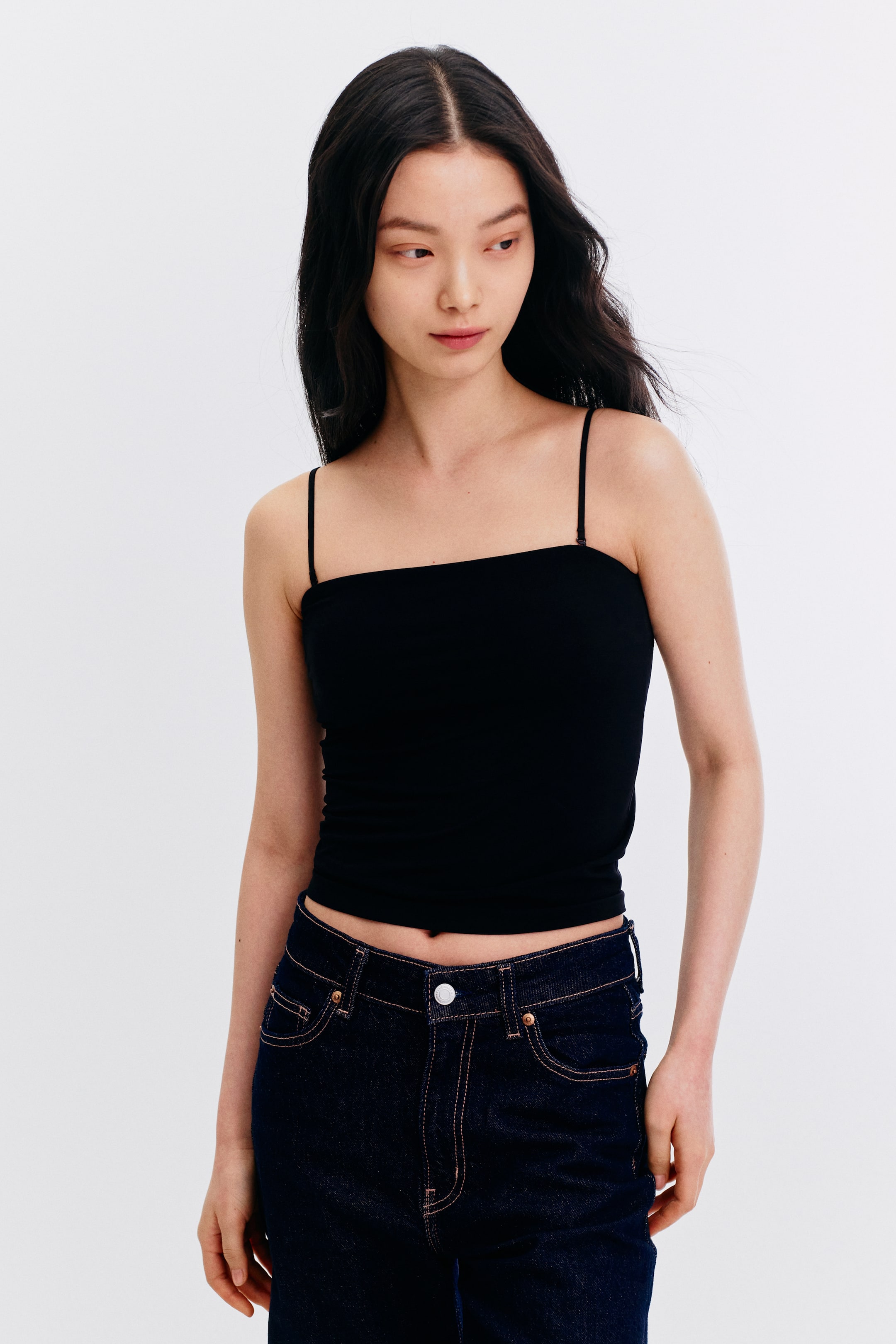 View larger image: The model is showcasing a black camisole top featuring a square neckline and slender spaghetti straps. The top is tailored to fit closely and is cropped, allowing a glimpse of the waistband of dark wash jeans.
