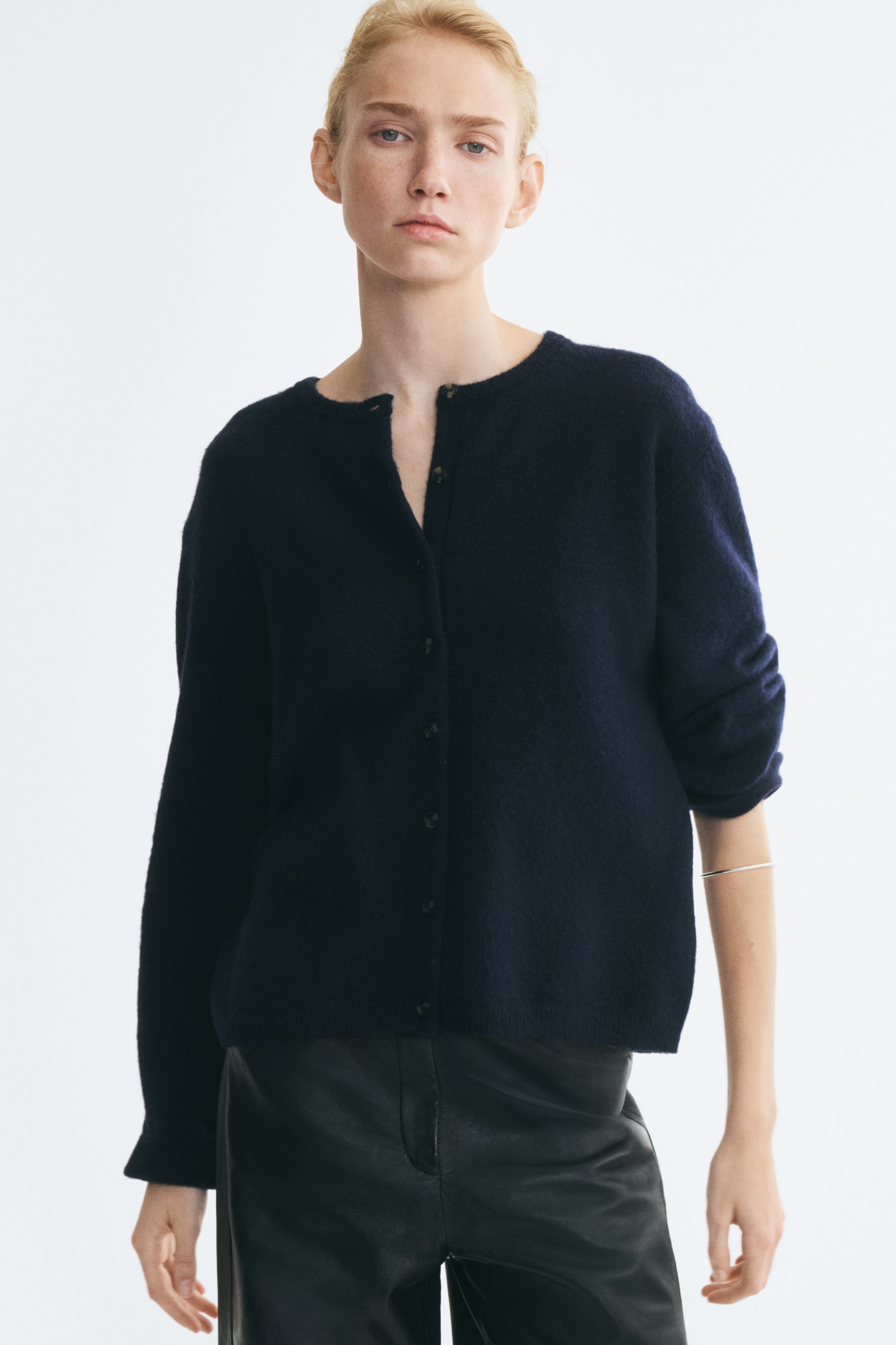 View larger image: A dark navy blue, loose-fit knitted cardigan is worn by the model, featuring a round neckline, button-down front, dropped shoulders, and long sleeves. The cardigan is styled with the top buttons left open and paired with black leather trousers.