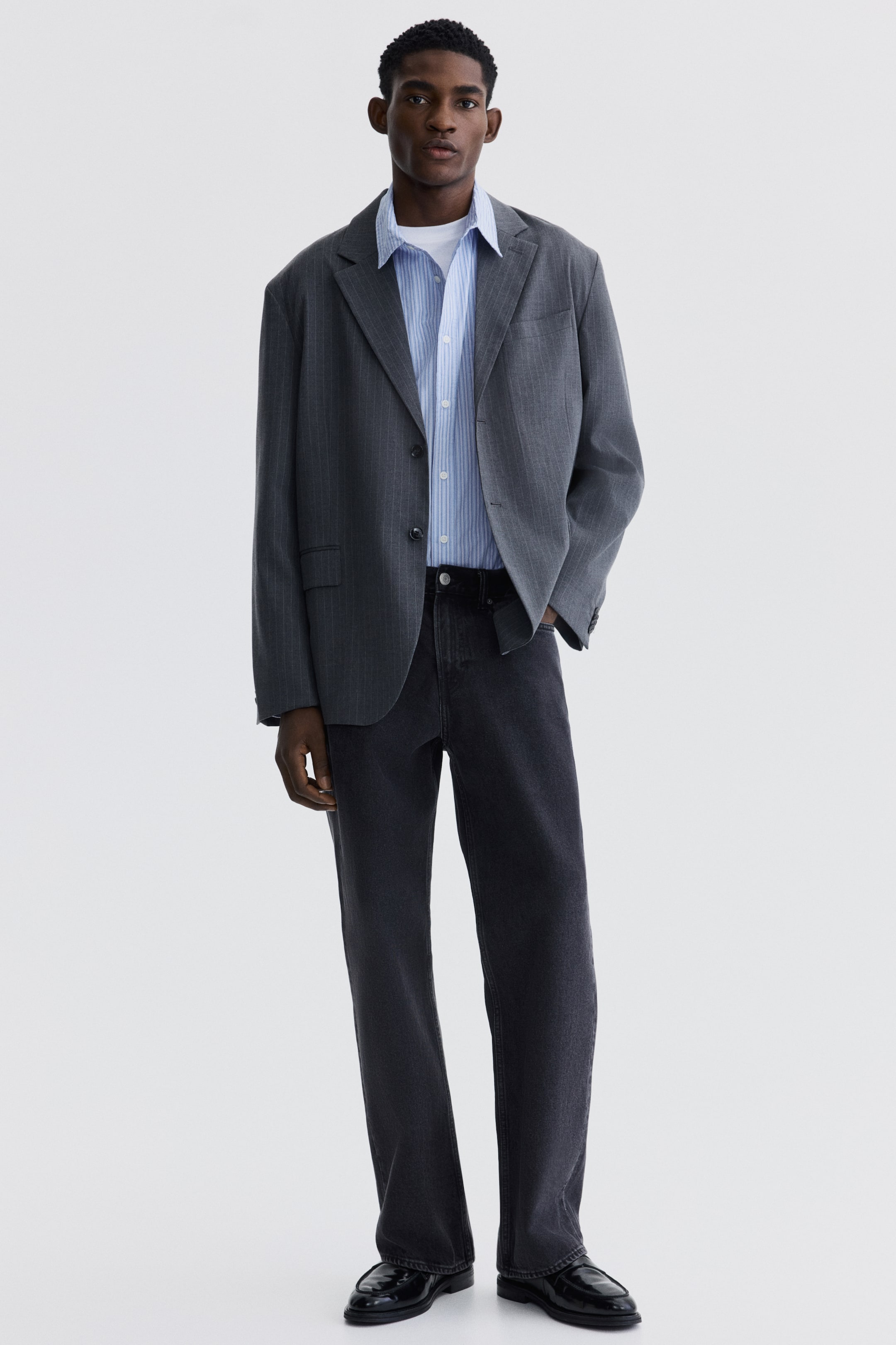 Büyük resmi görüntüle: Kombin, önden bakış açısıyla sunuluyor ve gri çizgili, çentikli yaka bir blazer ceket, açık mavi ve beyaz çizgili bir düğmeli gömlek üzerine giyilmiş. Gömlek, beyaz bir tişörtün üzerine katmanlanmış ve koyu gri düz paça kot pantolonlar siyah loafer ayakkabılarla tamamlanmış.