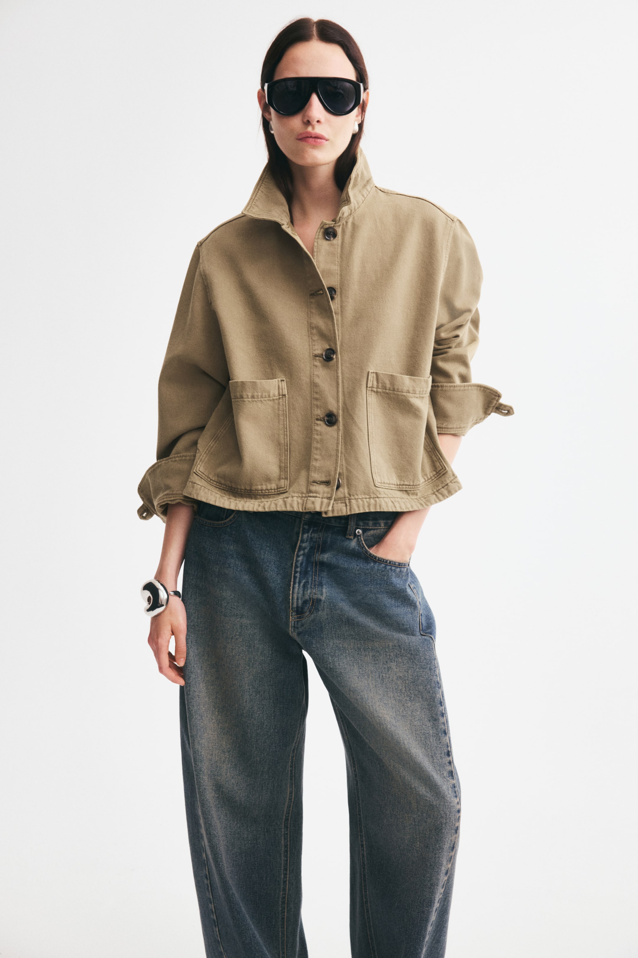 View larger image: A beige cotton denim jacket featuring a high collar, a button-down front, and large patch pockets is being worn by the model. The jacket has its long sleeves rolled up at the cuffs and is styled with blue denim jeans.
