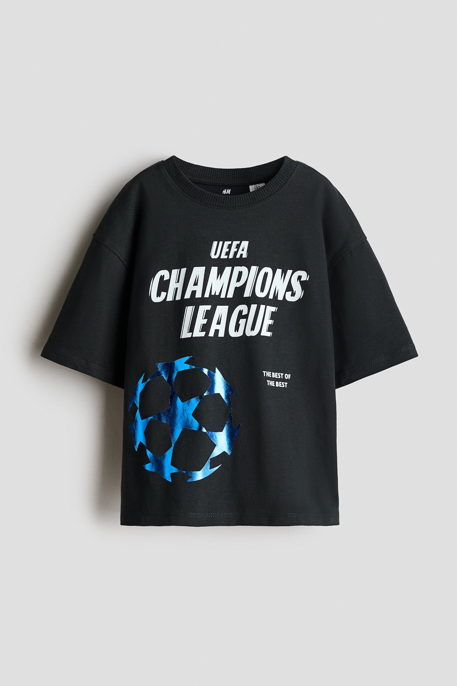 En oversized svart t-shirt i bomullsjersey visas framifrÃ¥n, med rund halsringning med ribbstickad kant och nedhasade axlar. Framsidan pryds av ett vitt tryck med texten âUEFA CHAMPIONS LEAGUEâ samt en glansig, metallisk blÃ¥ starball-grafik lÃ¤ngst ner till vÃ¤nster.