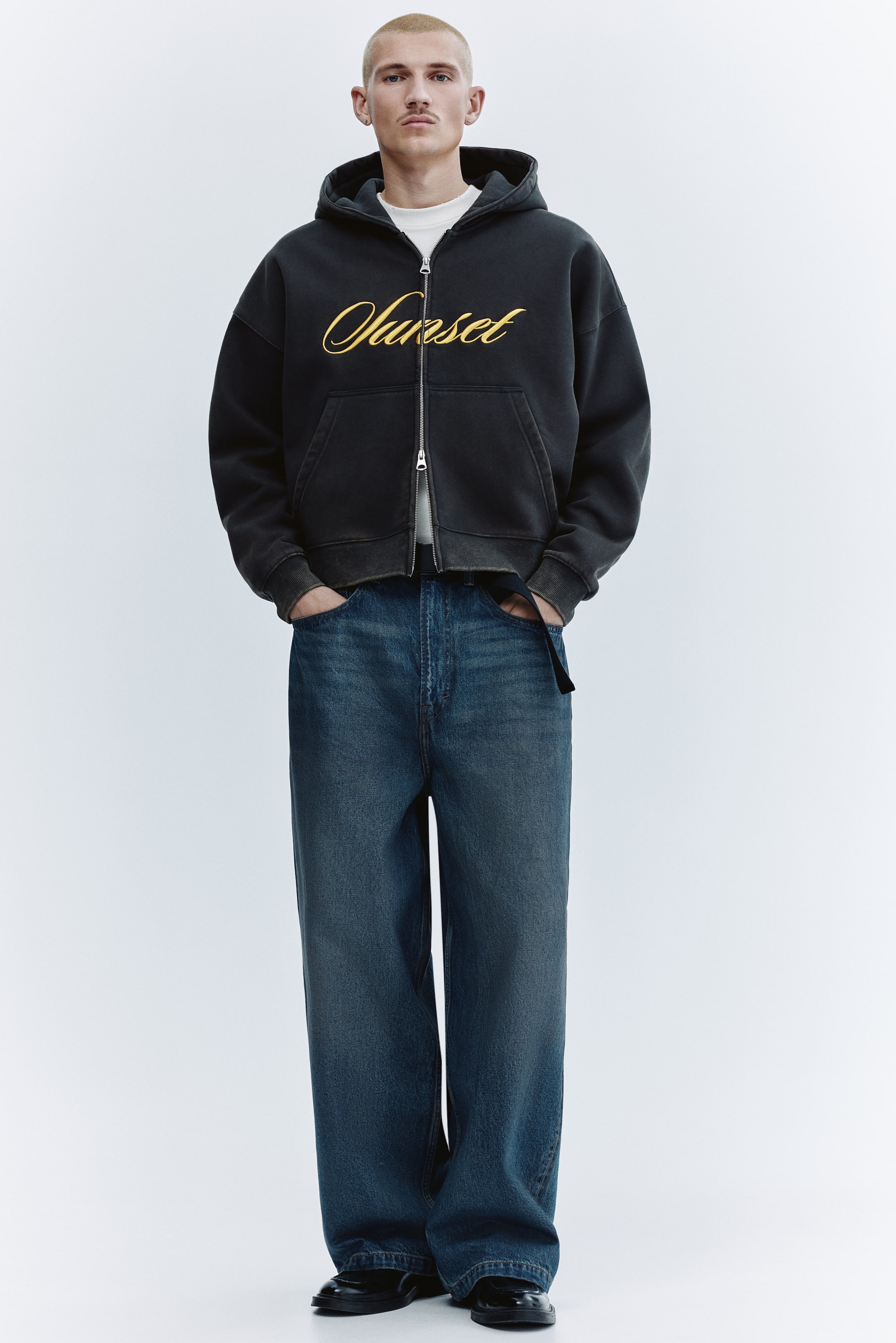 Ampliar la imagen: Una sudadera con cierre completo color gris oscuro presenta un bordado amarillo con la palabra "Sunset" en estilo script sobre el pecho, con un corte oversize y puños y dobladillo acanalados. Va superpuesta a una camiseta blanca y unos jeans de lavado oscuro de pierna ancha completan el look.