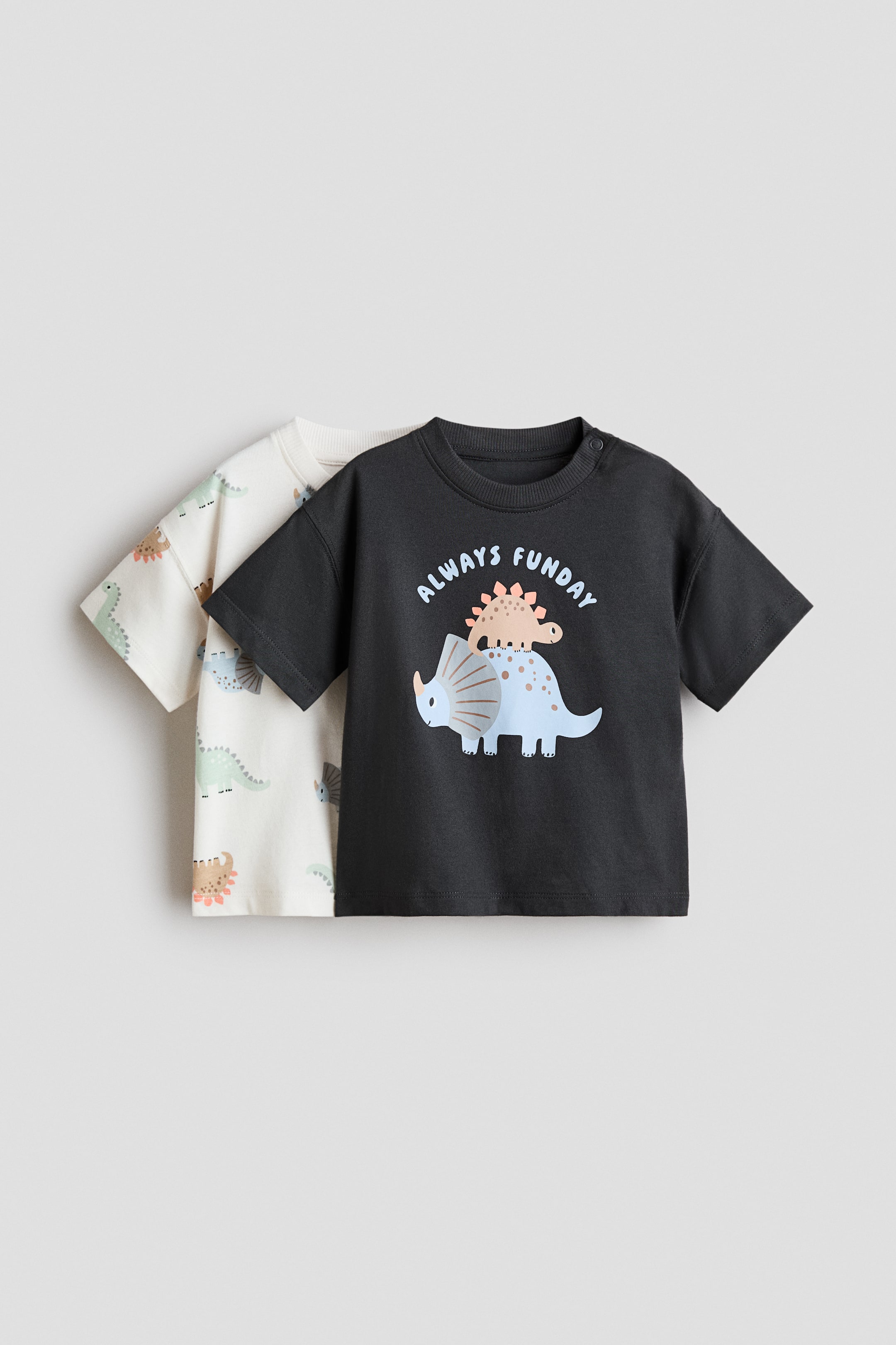 View larger image: Two short-sleeved t-shirts are layered, facing FRONT. The dark gray t-shirt in front is featuring a graphic of a light blue triceratops and a smaller brown stegosaurus, with 'ALWAYS FUNDAY' text, a ribbed crew neck, and two snap buttons on the left shoulder. The off-white t-shirt behind it is displaying an all-over print of various cartoon dinosaurs.