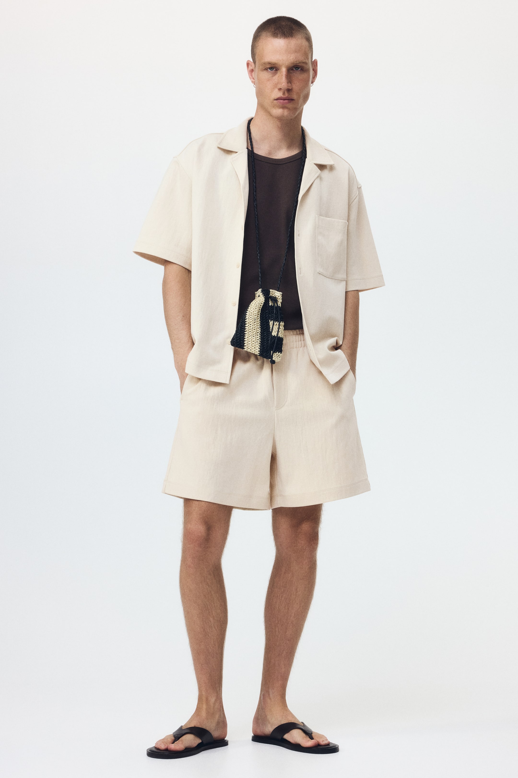 View larger image: A beige short-sleeve shirt with a camp collar and a single chest patch pocket is worn open over a dark brown t-shirt. Matching beige shorts with an elastic waistband and side pockets complete the relaxed, forward-facing ensemble.