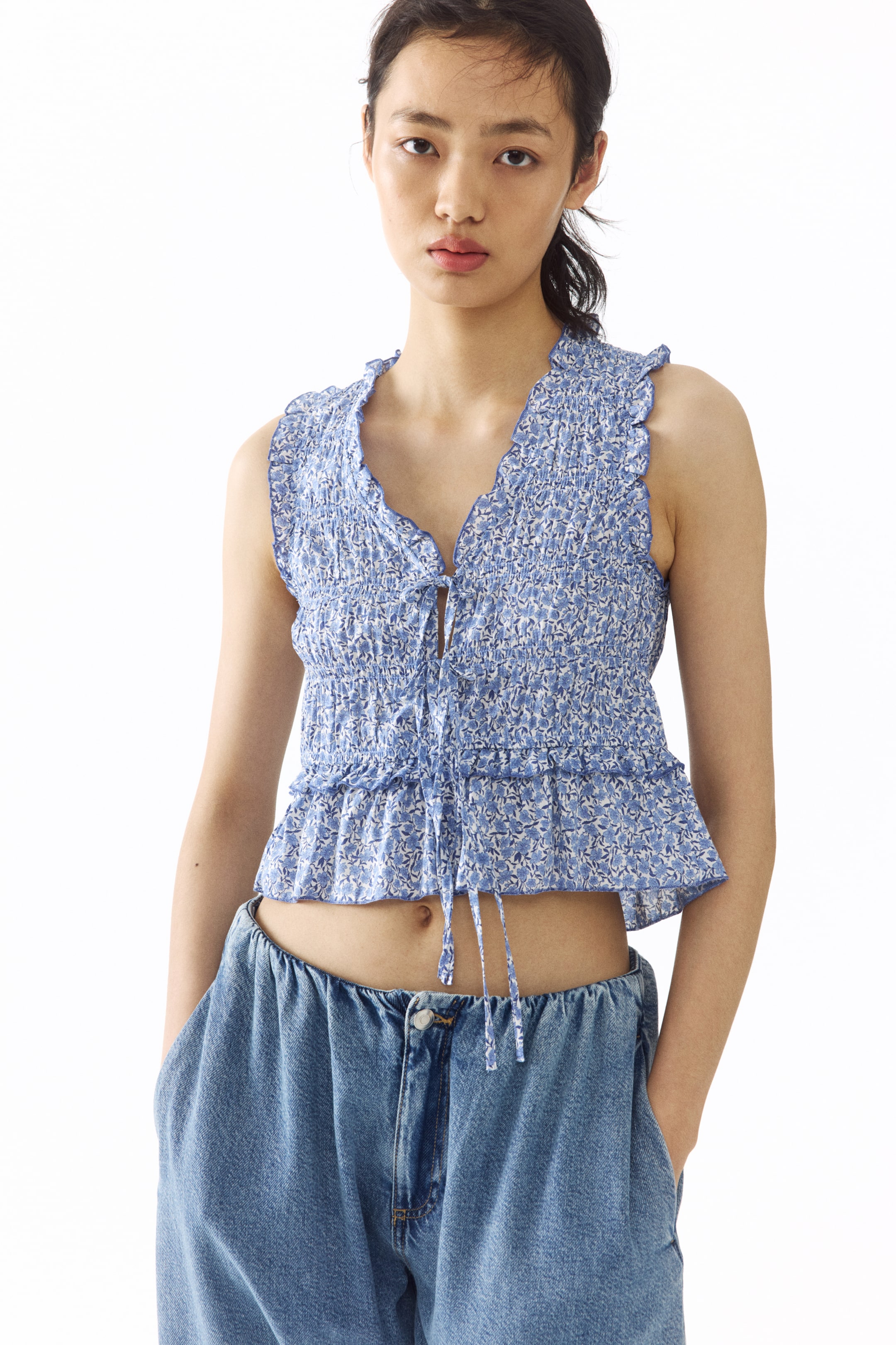 View larger image: This sleeveless top highlights a blue and white floral print, featuring a smocked bodice and a V-neckline with ruffled trim. Designed with a tie-front closure, it has a cropped length and a ruffled peplum hem, styled over light blue jeans.