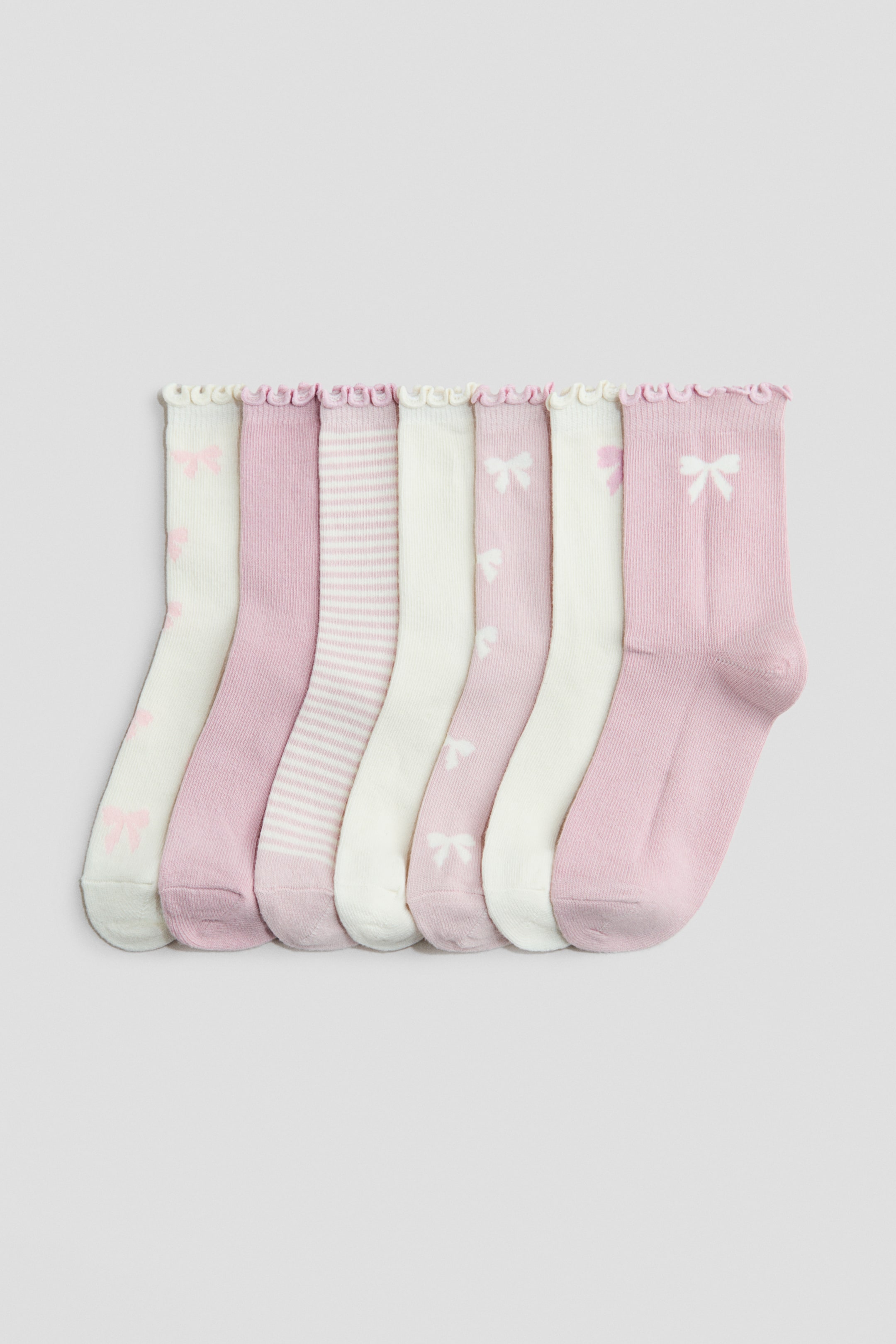 View larger image: Fine-knit socks with elasticated tops are standing upright in a row, featuring ruffled edges. The set of seven socks is showcasing alternating light pink and off-white colours with solid, bow-patterned, and horizontally striped designs.