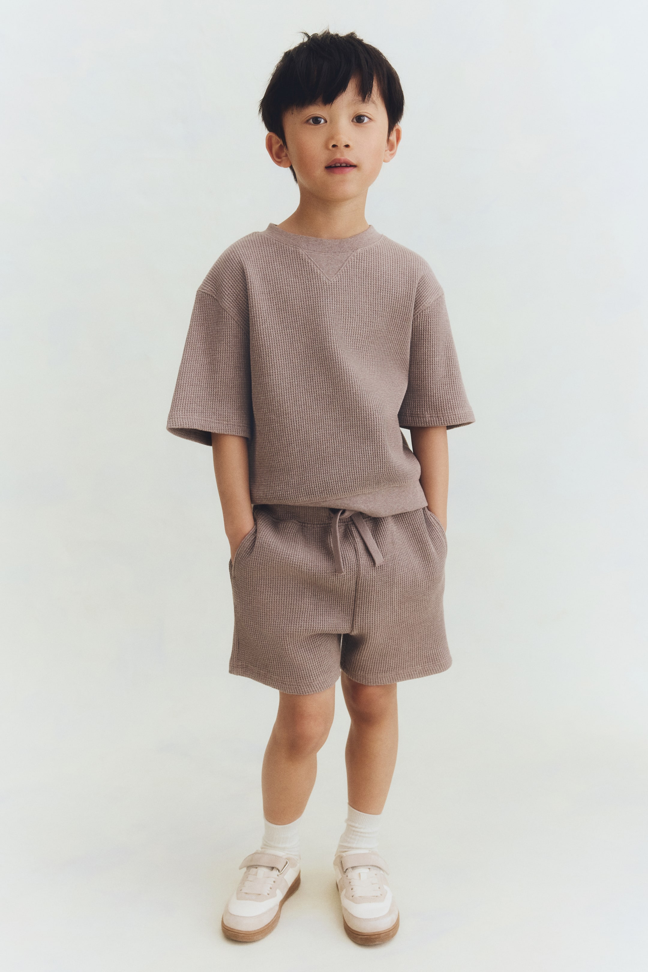 Agrandir l'image: Un ensemble t-shirt et short en jersey gaufré taupe est présenté de face. Le t-shirt est doté d'un col rond côtelé avec un détail en V en dessous, des épaules tombantes et des manches longueur coude. Le short est équipé d'une taille élastiquée avec cordon de serrage et de poches latérales.