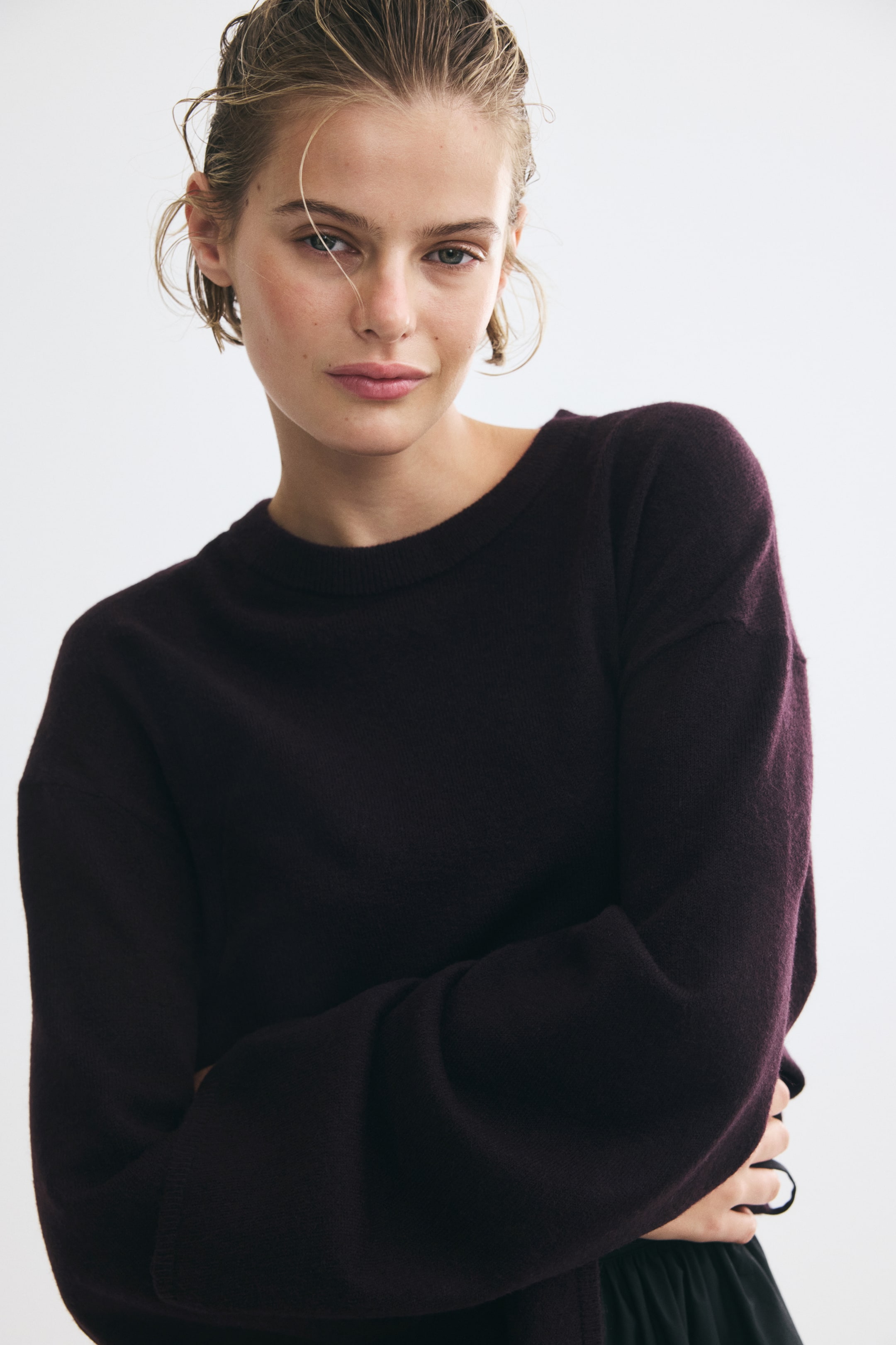 View larger image: A dark plum-colored knit sweater featuring a ribbed crew neckline and dropped shoulders is being worn by the model. The model is standing with her arms crossed, showcasing the long sleeves and soft texture of the garment.