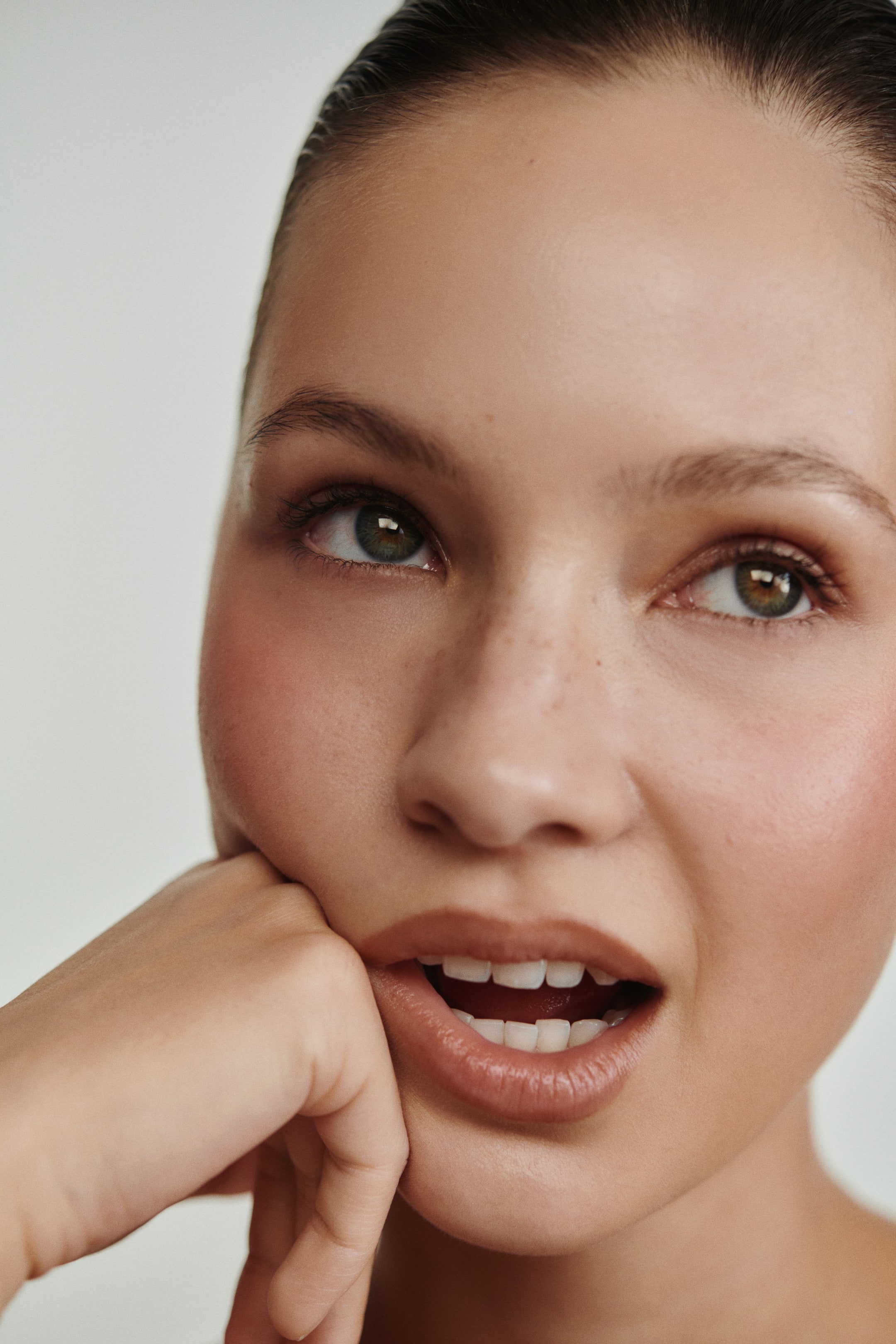 Größeres Bild anzeigen: Das Gesicht einer Person wird aus einem leicht aufwärts gerichteten Winkel präsentiert, wobei klare Haut mit sichtbaren Sommersprossen und rosigem Rouge auf den Wangen zu sehen ist. Definierte Augenbrauen rahmen Augen ein, die nach oben schauen, während die Lippen leicht geöffnet sind und einen nudefarbenen Lippenstift sowie Zähne zeigen. Eine Hand ruht an der unteren Kieferlinie.