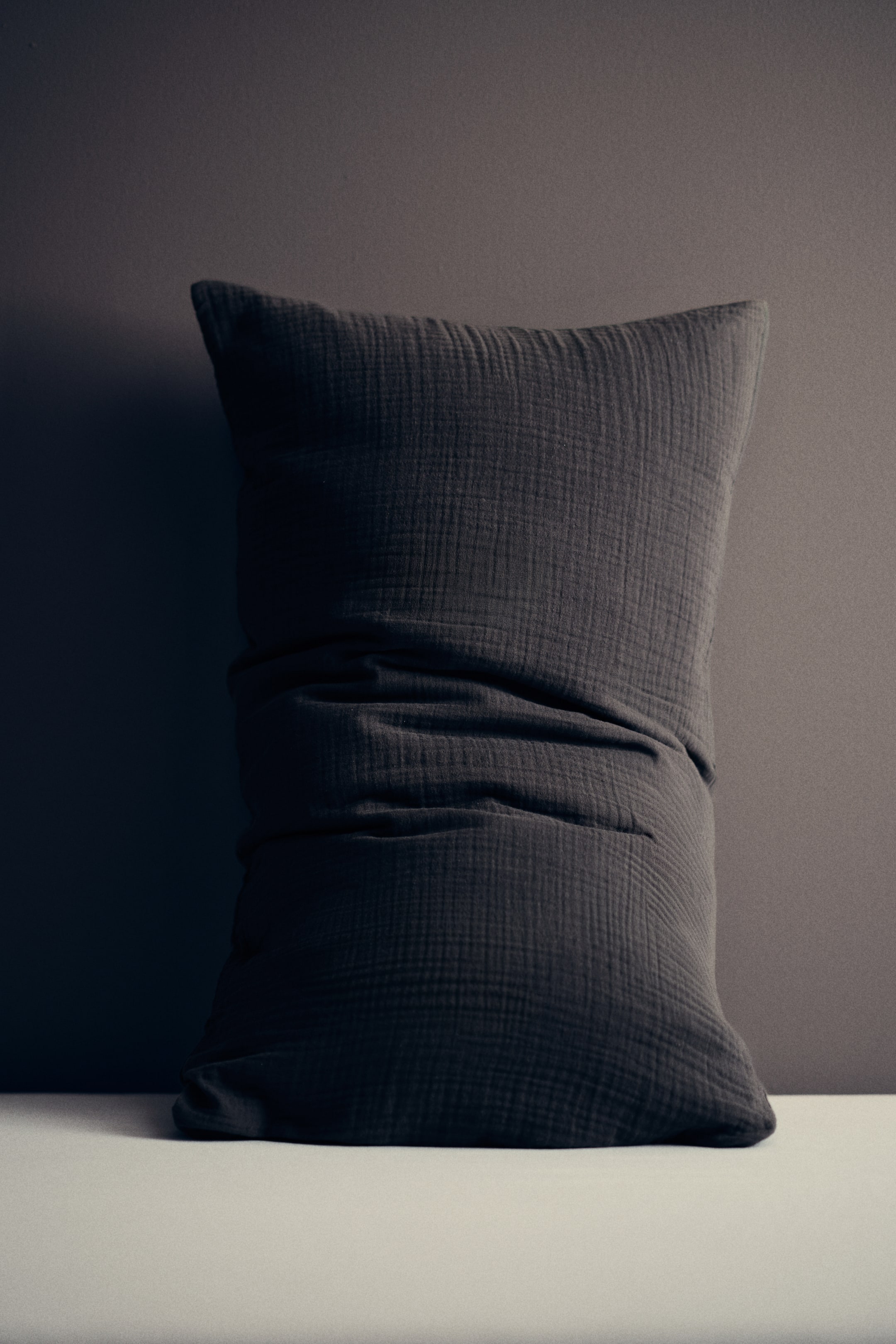 Größeres Bild anzeigen: Ein dunkelgraues rechteckiges Kissen steht aufrecht und weist eine geknitterte Stoffstruktur mit markanten Falten im unteren mittleren Bereich sowie sanft abgerundeten oberen Ecken auf.