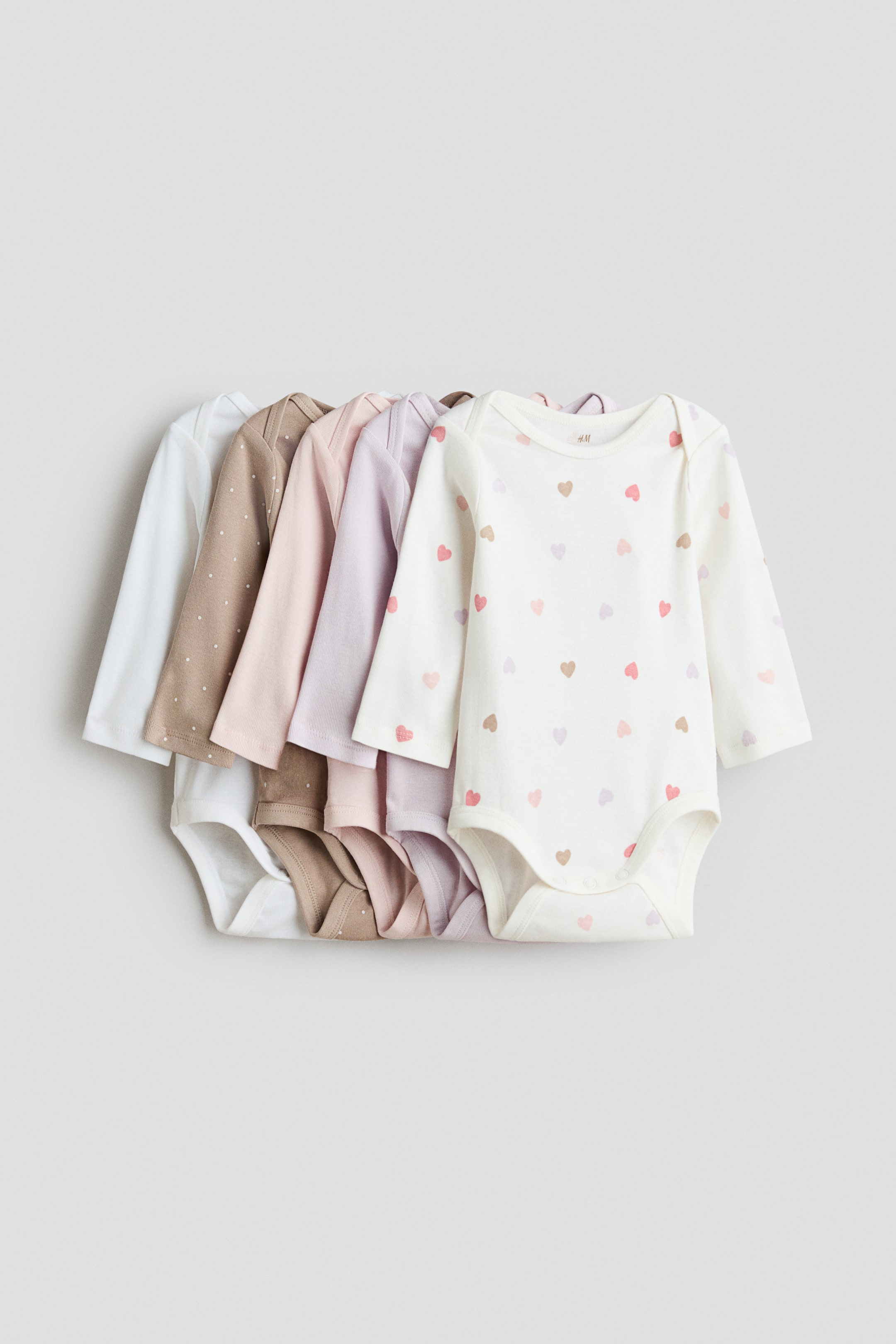 View larger image: Five long-sleeve baby bodysuits are displayed in a front-facing, overlapping row. The set features a cream-coloured bodysuit with a multi-coloured heart pattern on the front, followed by solid light purple, solid light pink, brown with white polka dots, and solid white options, all designed with envelope necklines and press-stud closures at the bottom.