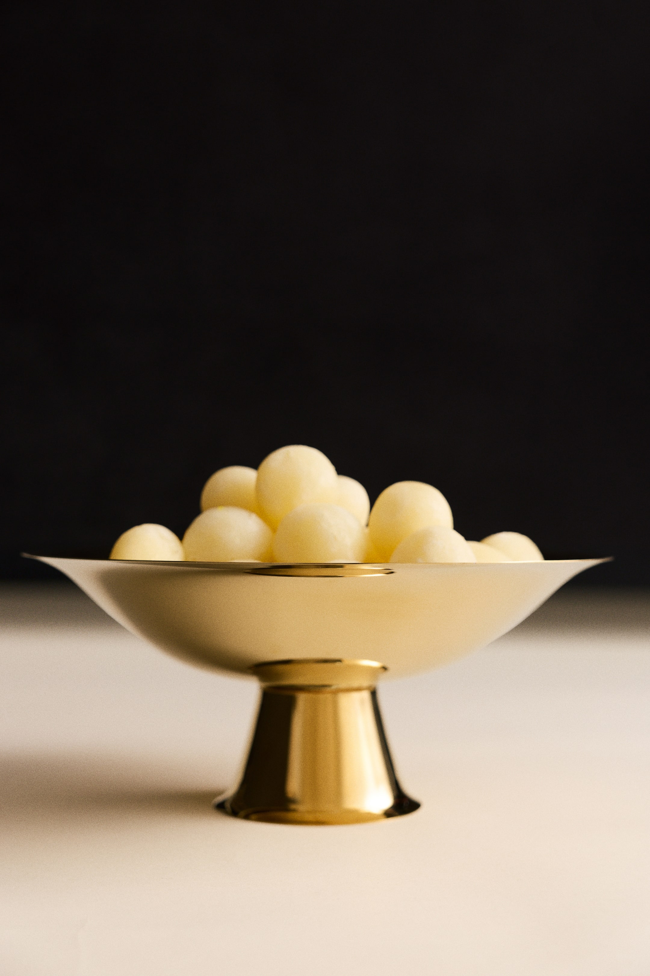 View larger image: A gold-toned stainless steel footed bowl, shown from a front angle, featuring a wide, shallow flared basin supported by a polished, tapered pedestal base. The bowl holds a mound of small, pale yellow spherical melon scoops.