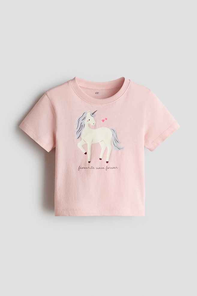 A pink short-sleeved t-shirt features a cream-coloured unicorn graphic with a grey mane and tail on its front, accompanied by two small pink hearts near its head and the text 'favourite since forever' printed below. - 1