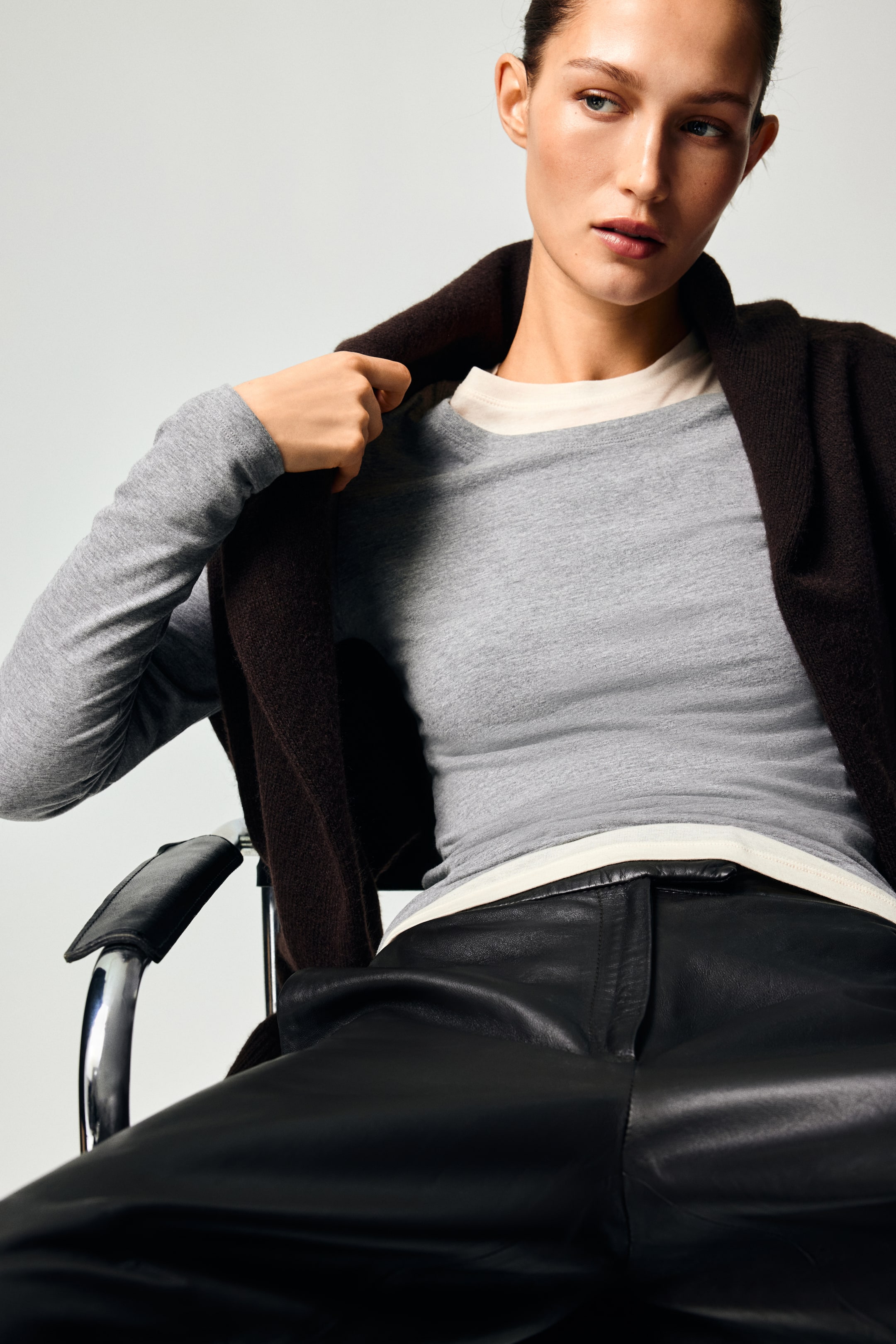 View larger image: A light-grey, long-sleeved crew-neck top fits closely, layered over an off-white t-shirt, with a dark-brown knitted cardigan draped over the shoulders. Black leather trousers feature a smooth texture and a visible fly-front closure.