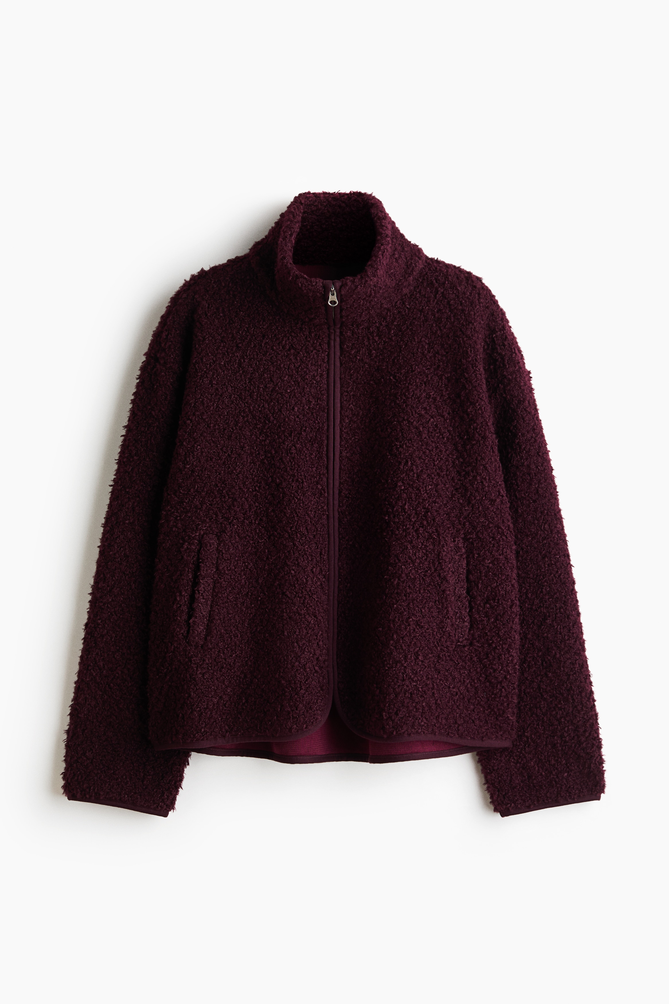 A dark burgundy teddy jacket with a stand collar, front zipper, and dropped shoulders is worn by the model. The jacket is styled with light-wash denim jeans.