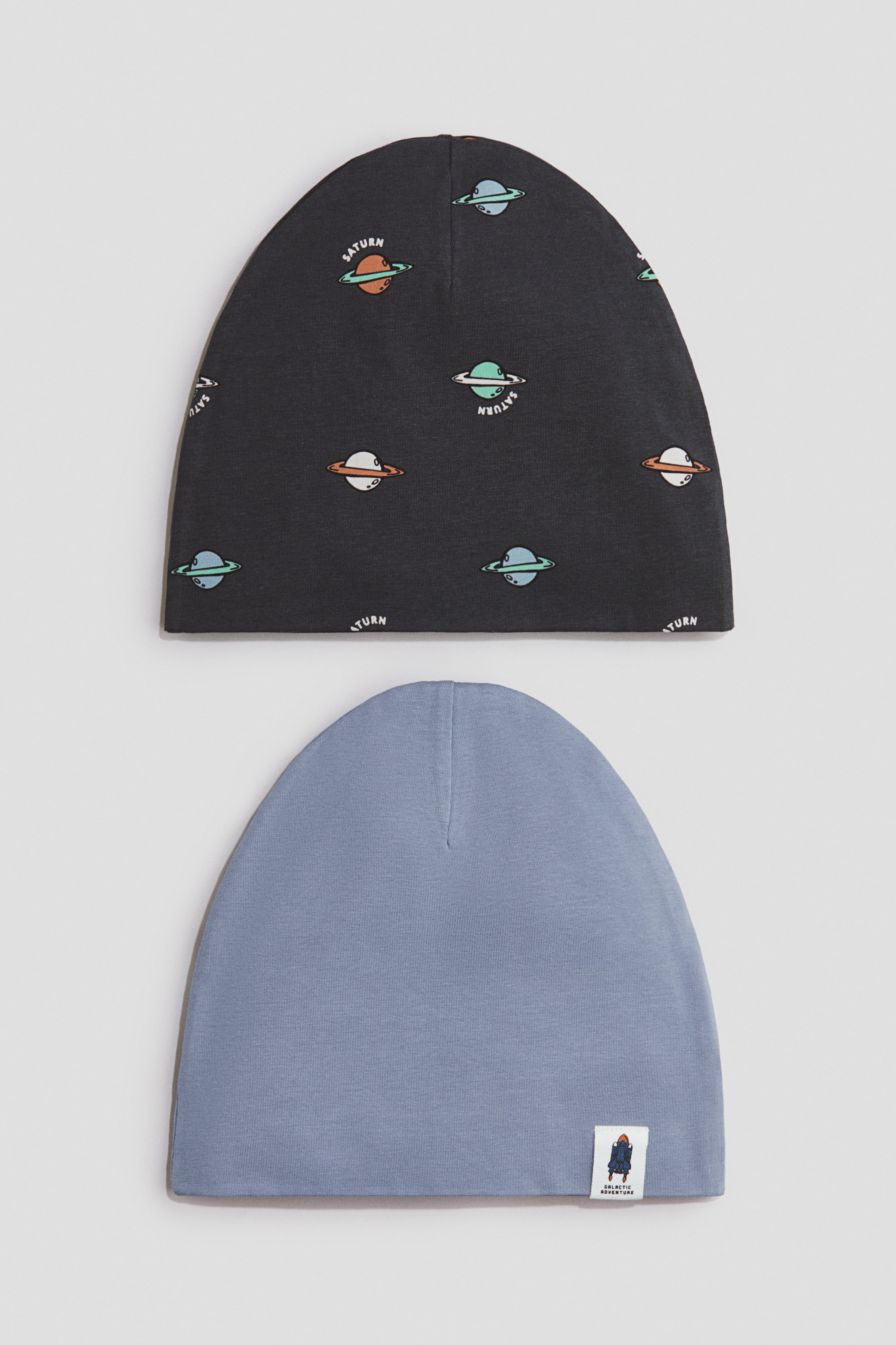 View larger image: A set of two beanies is displayed, comprising a dark grey beanie adorned with a repeating print of colourful cartoon planets with rings and the text "Saturn", and a light blue beanie showcasing a small fabric tag with a rocket graphic and the text "Galactic Adventure" on its lower right edge.