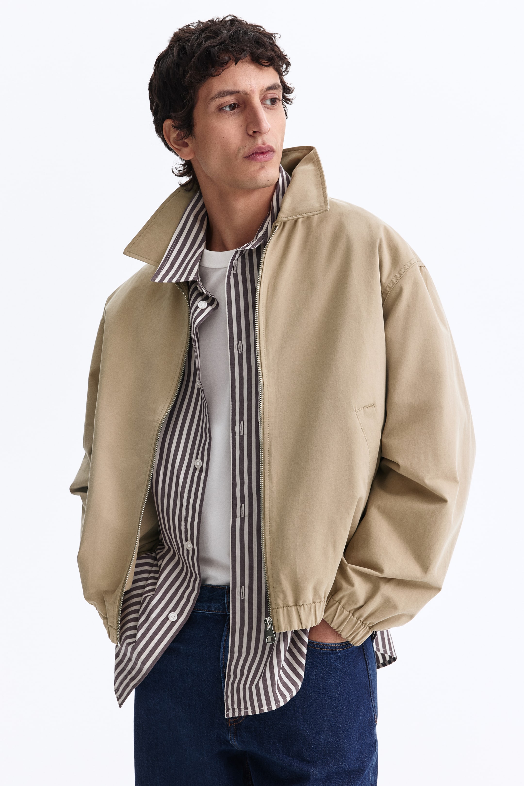 View larger image: A beige jacket is shown from a slightly angled front view, worn open to reveal a layered shirt underneath. It features a full-zip closure with a silver zipper, a pointed collar, long sleeves with elastic cuffs, and an elastic hem for a relaxed fit.
