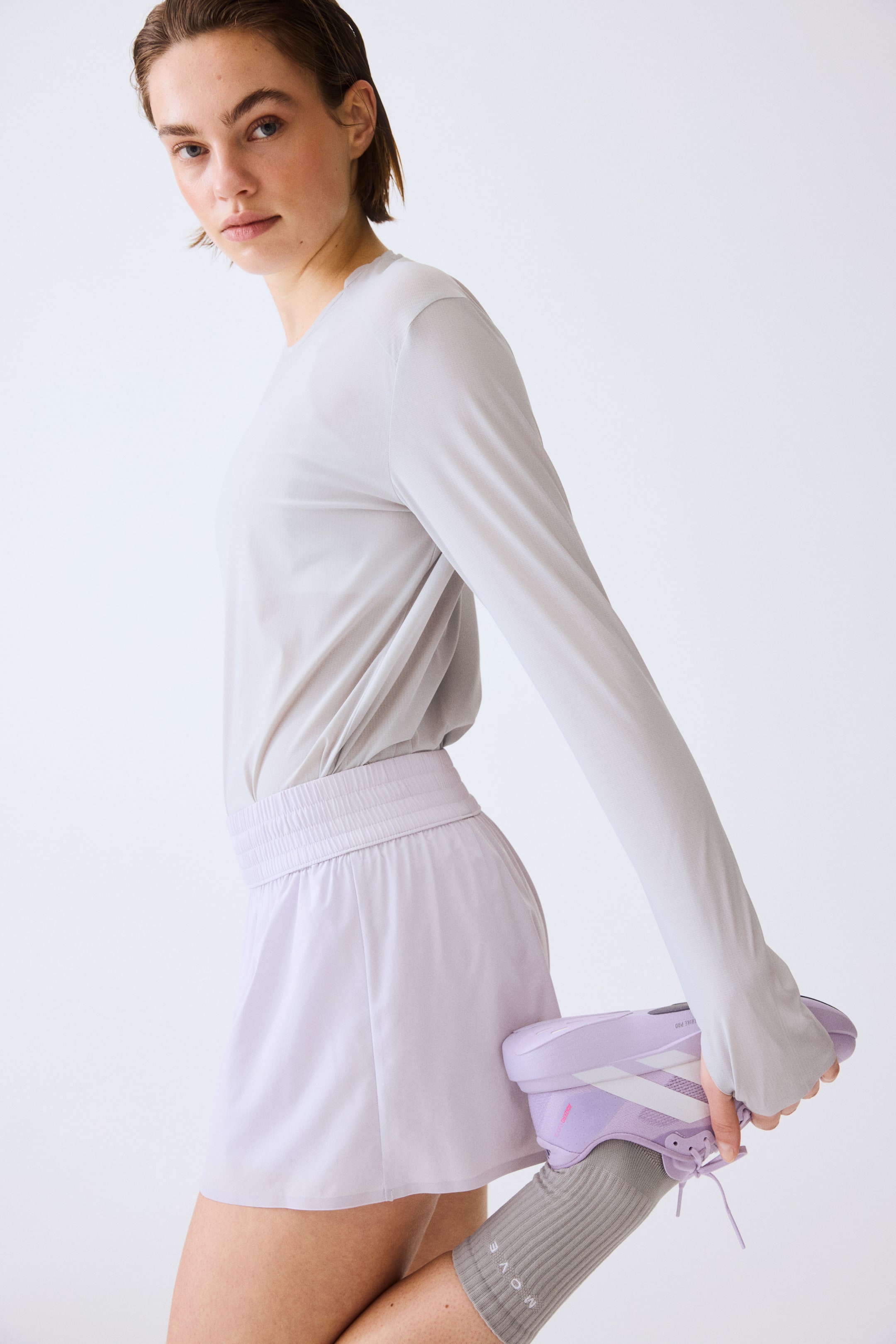 Visualizza immagine più grande: Pantaloncini sportivi a vestibilità regolare in lavanda chiaro, con elastico arricciato in vita, indossati a strati sopra pantaloncini da ciclismo in jersey. La modella si sta allungando, tenendo un piede, rivelando i pantaloncini da ciclismo in jersey che si estendono sotto l'orlo dei pantaloncini esterni.