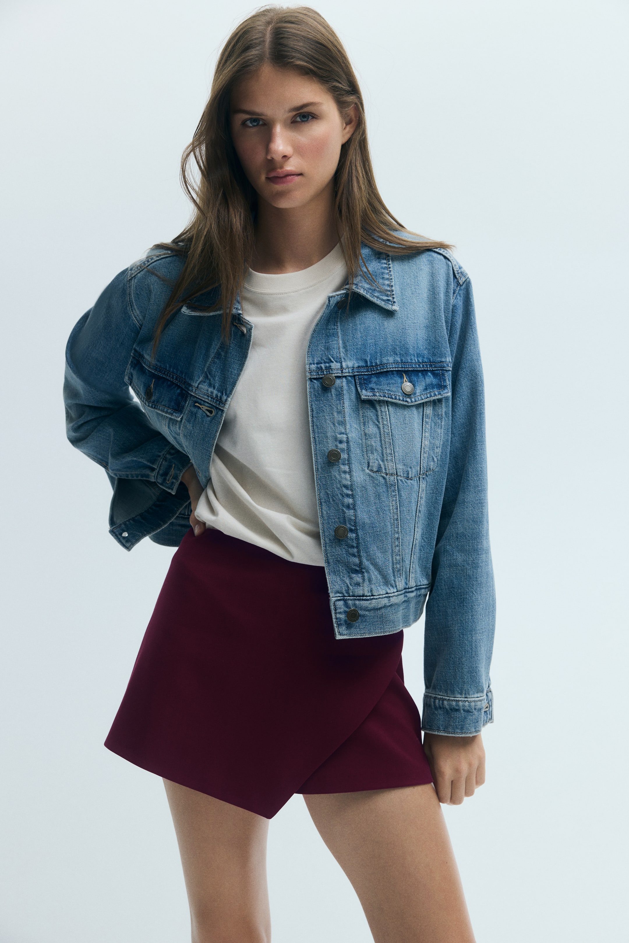 Ampliar la imagen: La modelo lleva puesto un skort envolvente color borgoña con un panel frontal asimétrico. El skort se combina con una playera blanca metida por dentro y una chaqueta de mezclilla azul.