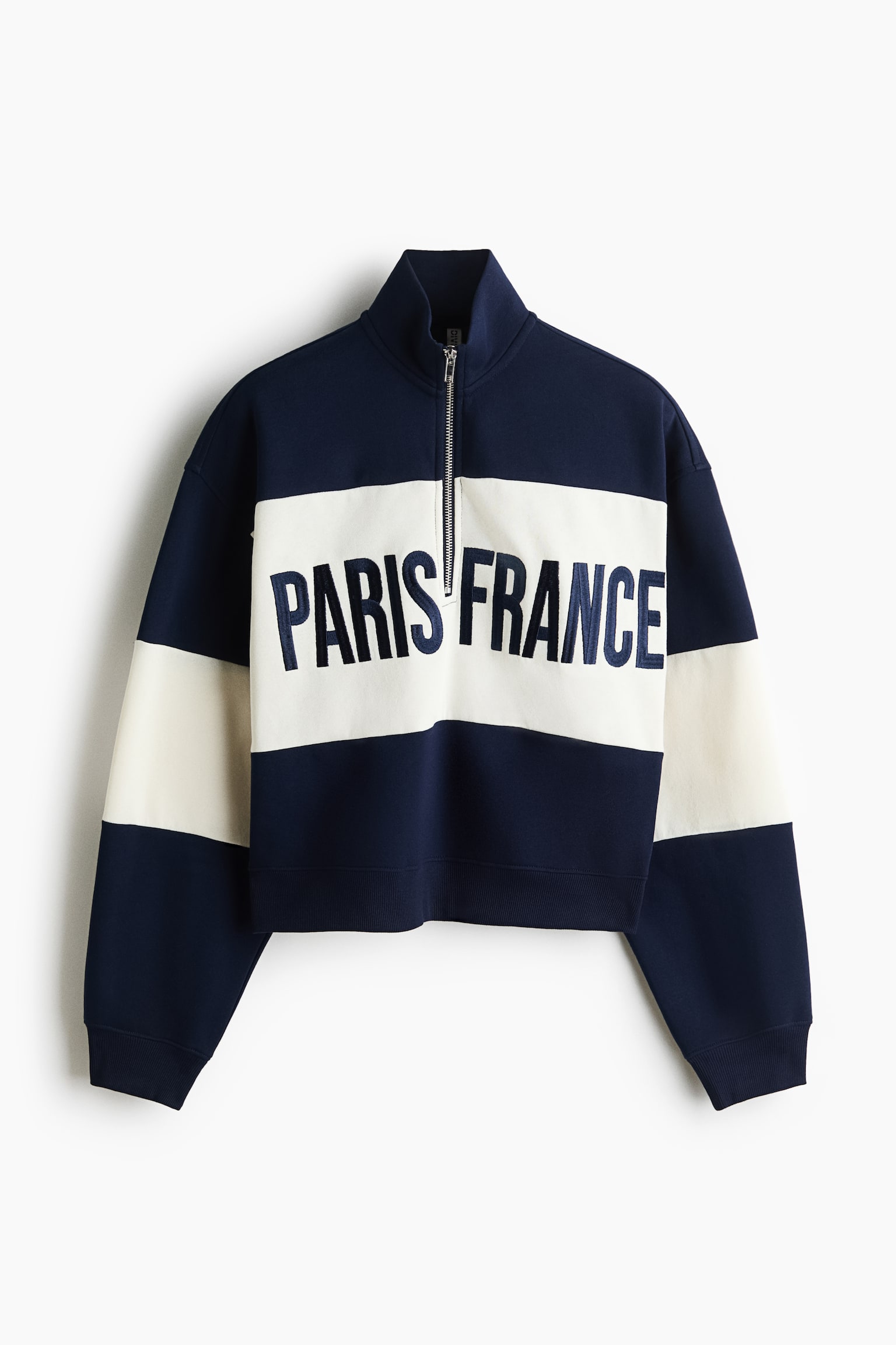 A navy blue cropped sweatshirt is shown from the front, featuring a high mock neck with a silver half-zip fastening. A wide, off-white horizontal stripe runs across the chest and sleeves, displaying the embroidered navy text "PARIS FRANCE" across the front above a ribbed hem.