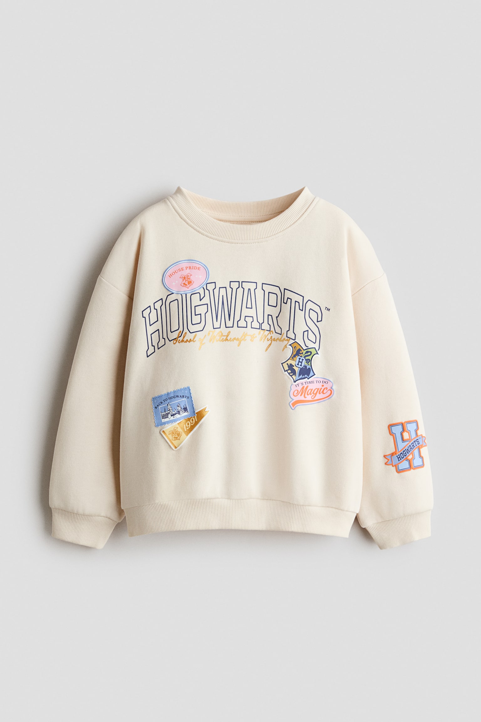 A cream-coloured sweatshirt is shown from the front, featuring a round neckline, dropped shoulders, and ribbed trim at the collar, cuffs, and hem. The chest displays a printed blue "HOGWARTS" graphic surrounded by various colourful school-themed patch designs, with an additional orange "H" graphic printed on the lower left sleeve.