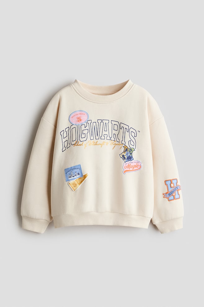En cremefarvet sweatshirt ses forfra med rund hals, sÃ¦nkede skuldre og ribkanter ved krave, manchetter og nederkant. PÃ¥ brystet ses et trykt blÃ¥t "HOGWARTS"-motiv omgivet af forskellige farverige skoleinspirerede patch-designs, og pÃ¥ den nederste venstre Ã¦rme er der desuden trykt et orange "H"-motiv. - 1