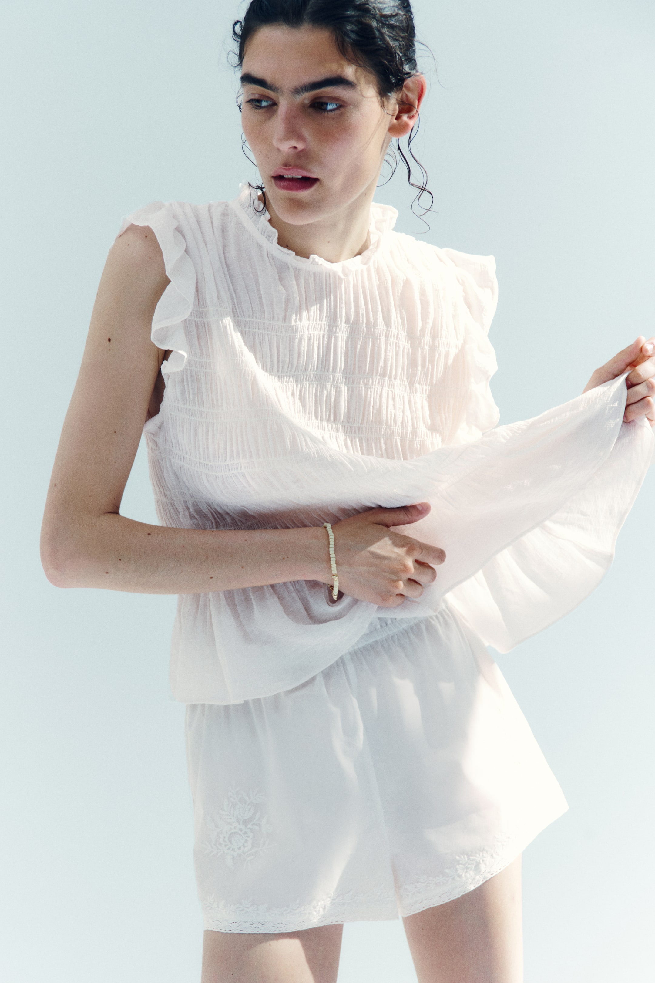 Grotere afbeelding bekijken: Een witte gesmokte peplumtop, met een gerimpelde hoge hals en gerimpelde kapmouwen, wordt gedragen door het model. Het model houdt de uitlopende peplumzoom zachtjes vast en de top is gestyled met bijpassende witte shorts.