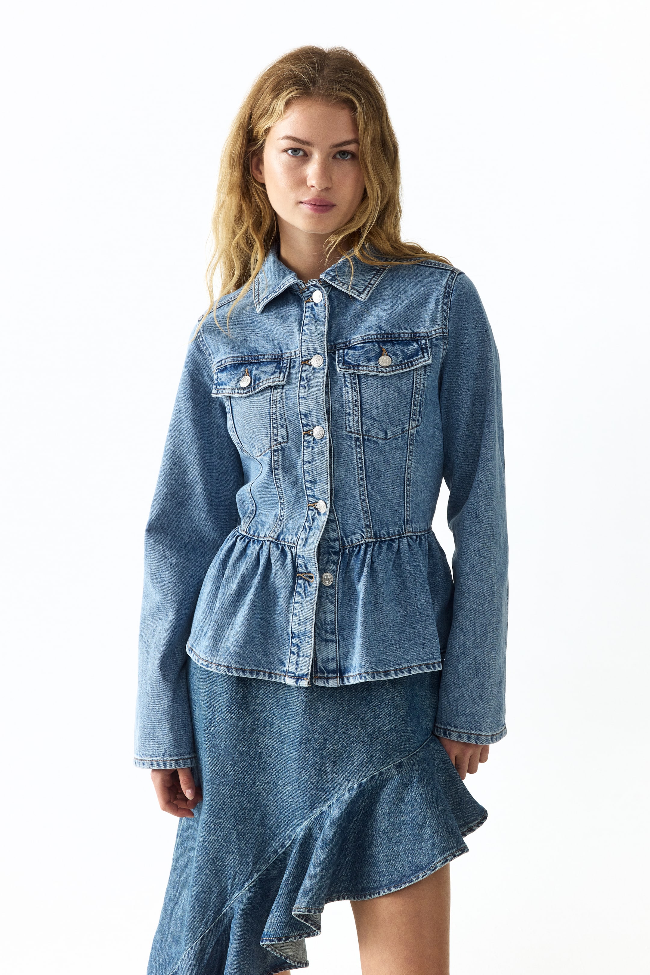 View larger image: A light blue wash denim peplum jacket featuring a pointed collar, a button-up front with silver-toned buttons, two chest flap pockets, and a fitted waist flowing into a gathered peplum hem. It's worn front-facing over a matching denim skirt showcasing an asymmetrical ruffled hem.