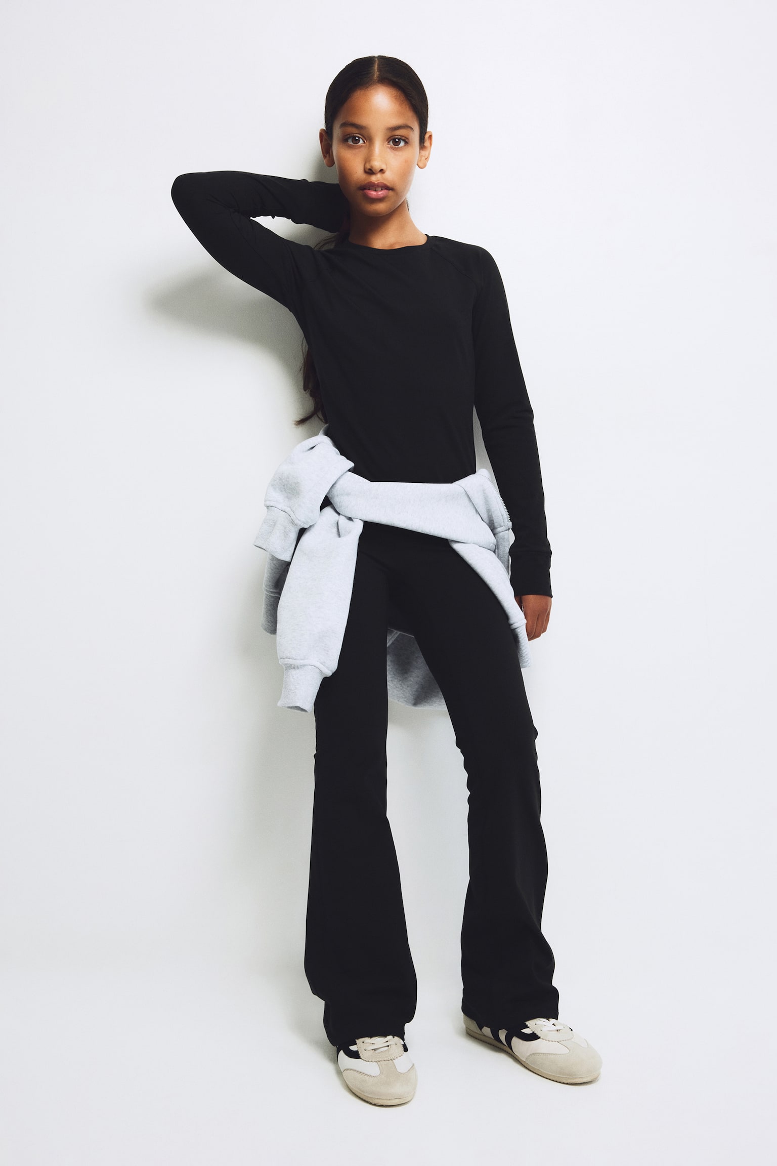 A front-facing view of a matching black set featuring a slim-fit, crew-neck long-sleeve top and flared trousers. A light grey sweatshirt is tied around the waist, layered over the fitted silhouette. - 1