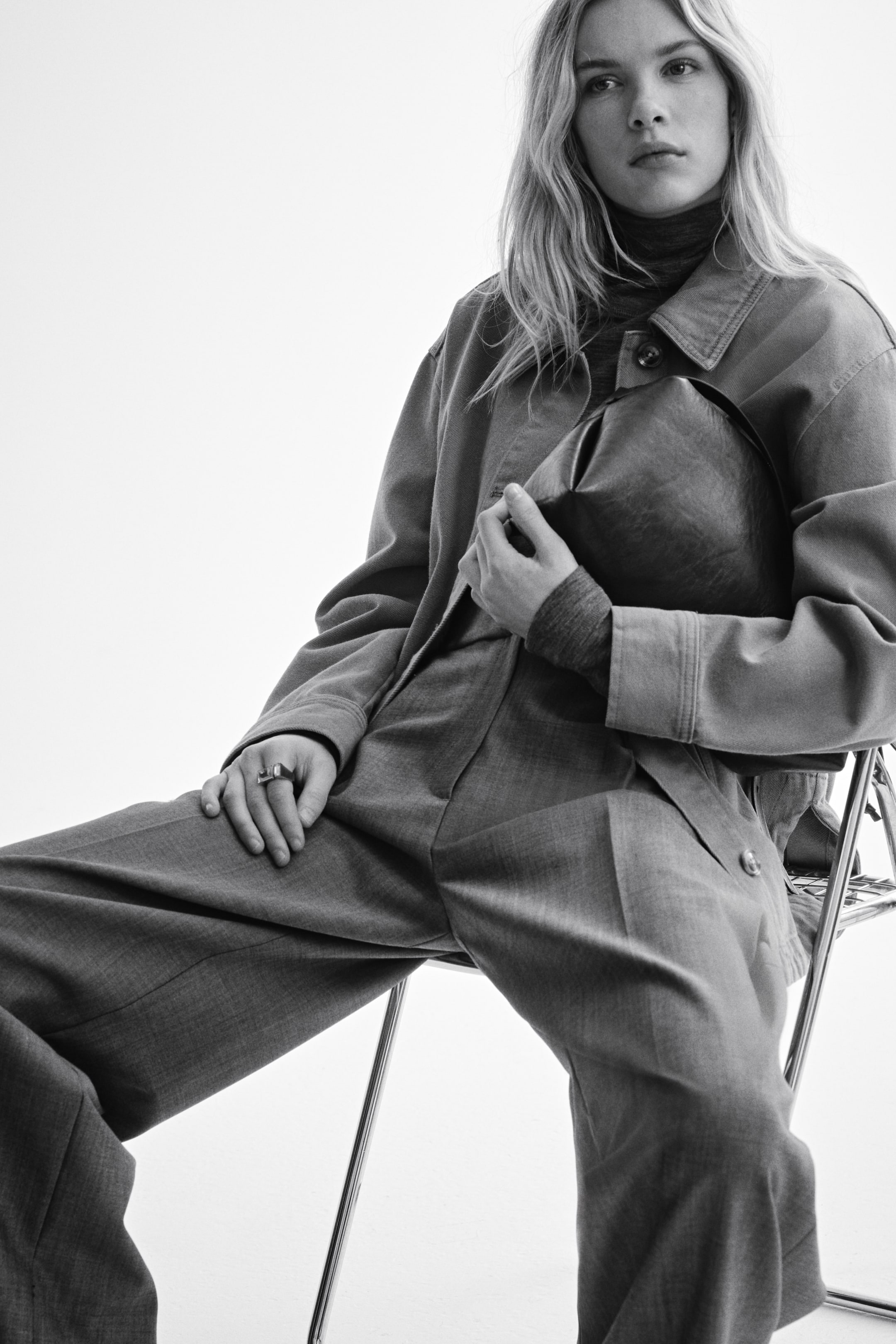 View larger image: A mid-gray oversized jacket with a classic collar and visible button closures is layered over a dark ribbed turtleneck. Mid-gray wide-leg pants with a pronounced front crease are worn, and a dark, soft, unstructured bag is held at the front.