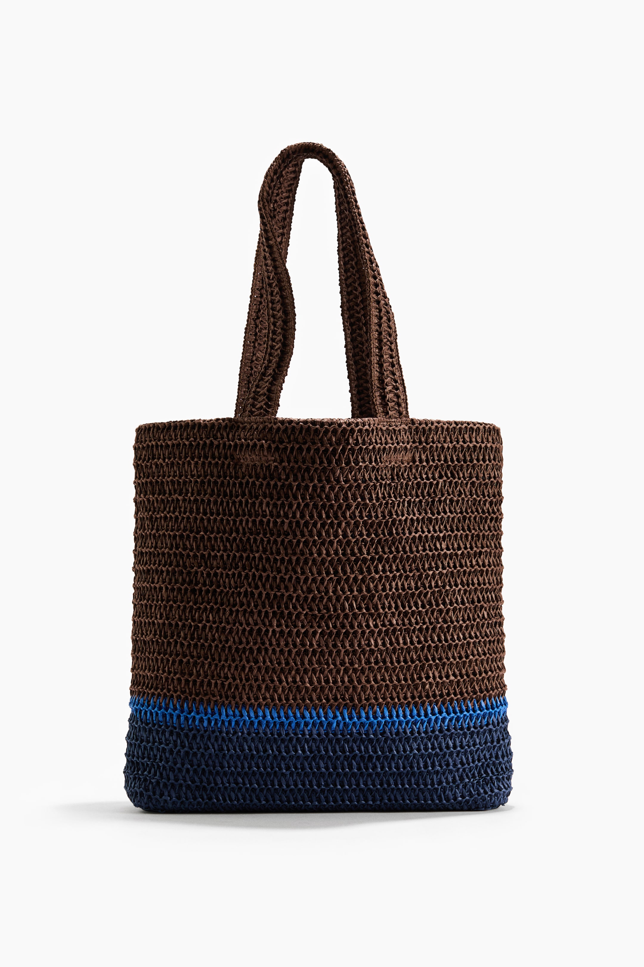 Größeres Bild anzeigen: Eine Tote Bag ist von vorne zu sehen, gefertigt aus geflochtenem Papierstroh. Sie verfügt über zwei geflochtene Tragegriffe, einen braunen oberen Teil, einen blauen horizontalen Streifen und einen dunkelblauen unteren Abschnitt.