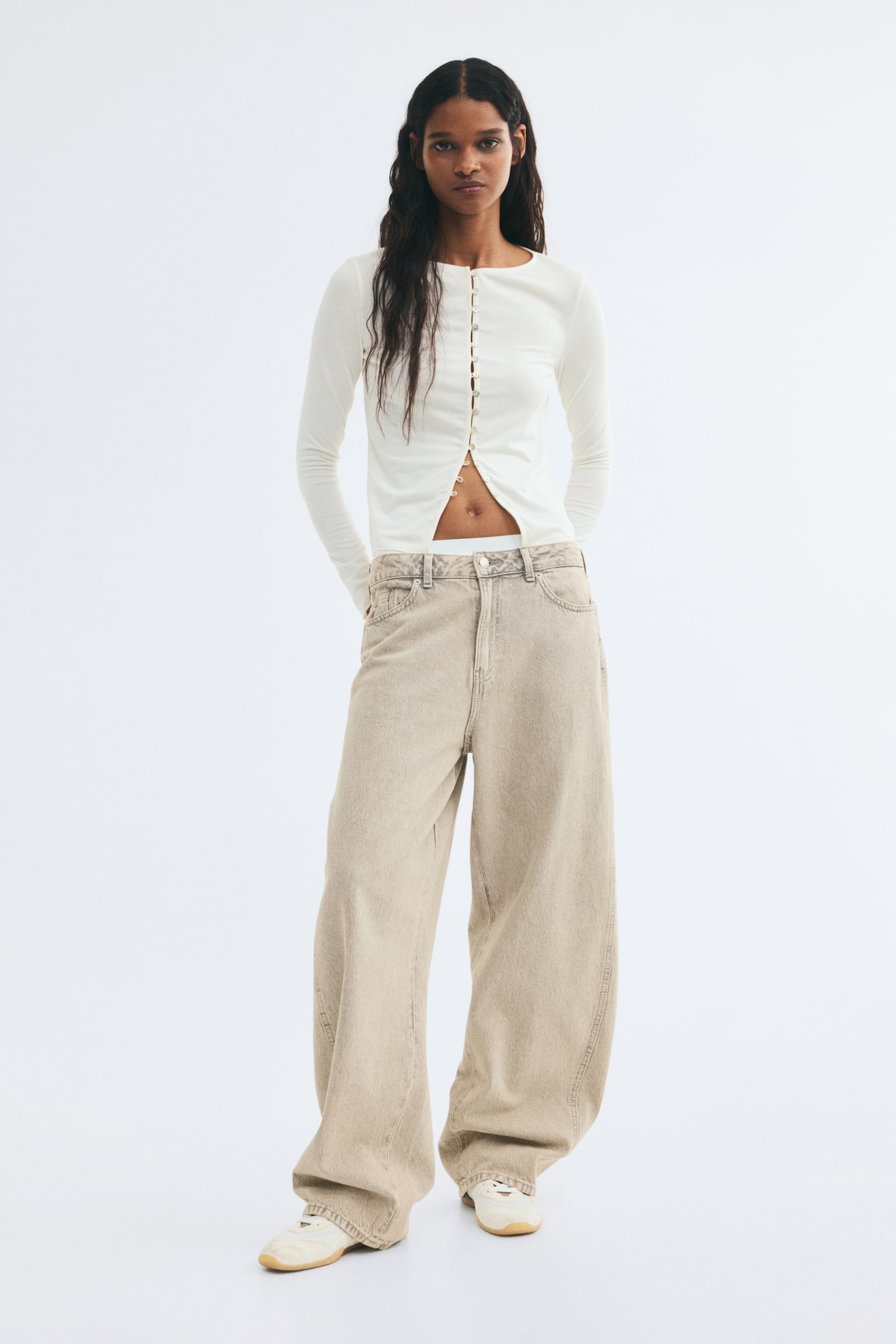A cream-coloured, fitted fine-knit top is being worn by the model, featuring a round neckline, long sleeves, and button-and-loop fastenings down the front. The top is left partially unbuttoned at the lower hem to reveal the midriff and is styled with wide-leg beige jeans. - 1