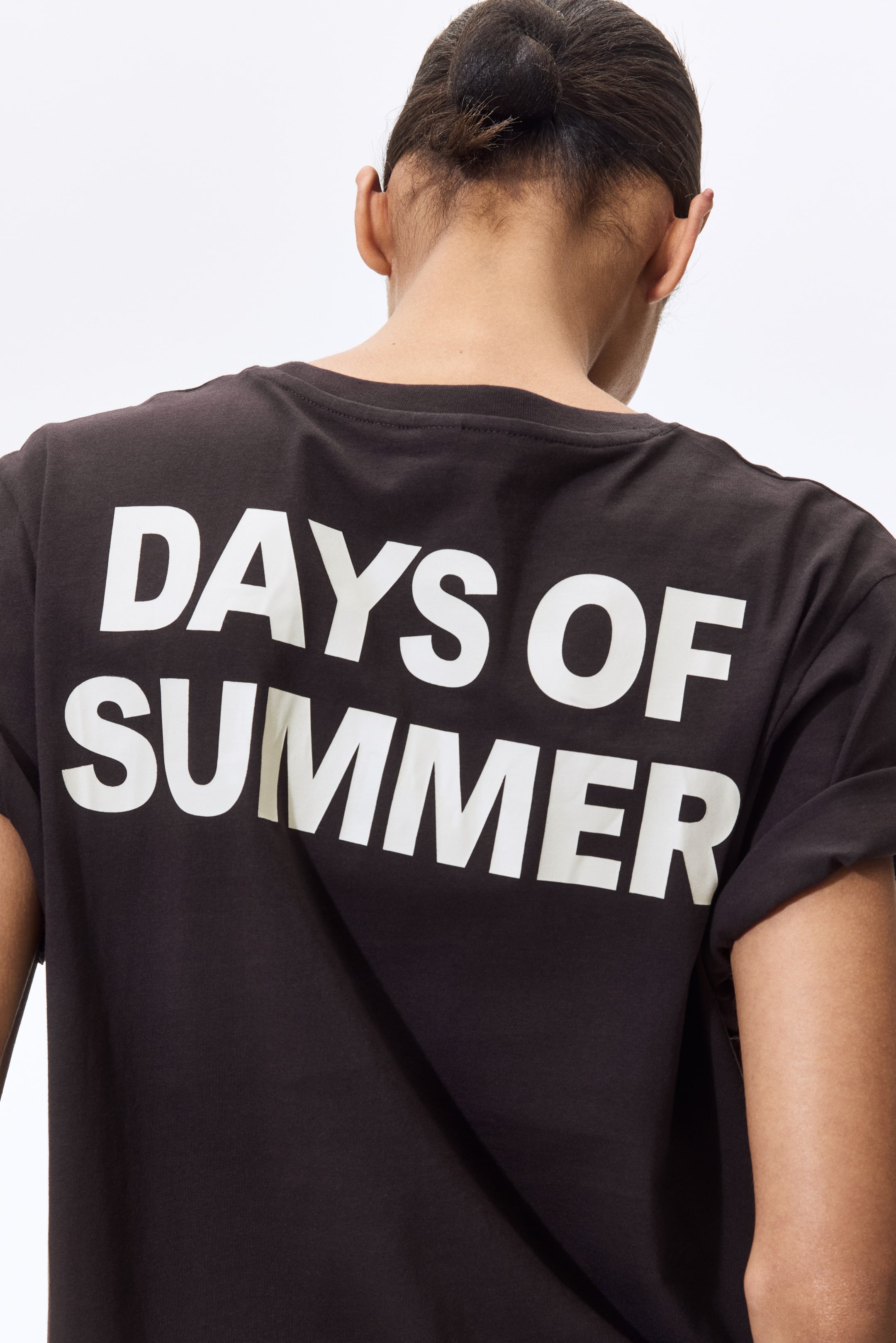 View larger image: A dark grey t-shirt is being worn, viewed from the back, displaying "DAYS OF SUMMER" in large white letters across the upper back. It features a crew neck, and its short sleeves are rolled up.