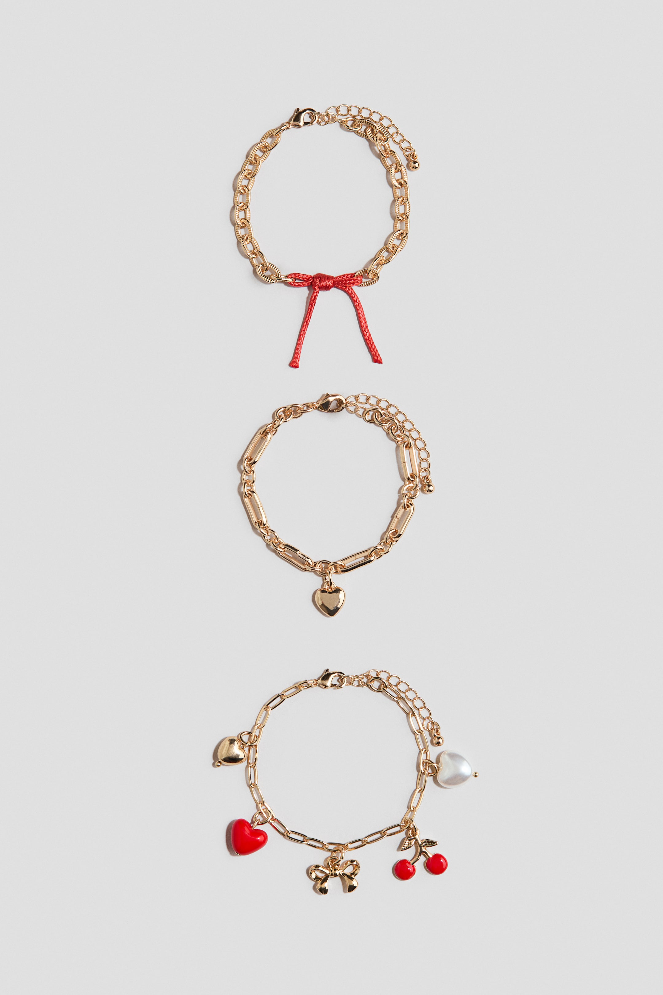 View larger image: Three gold-toned chain bracelets are lying flat, each featuring distinct charms. The top bracelet is displaying a red string bow, the middle bracelet is showcasing a gold heart charm, and the bottom bracelet is presenting multiple charms including red hearts, red cherries, a gold bow, and a white pearl-like heart.