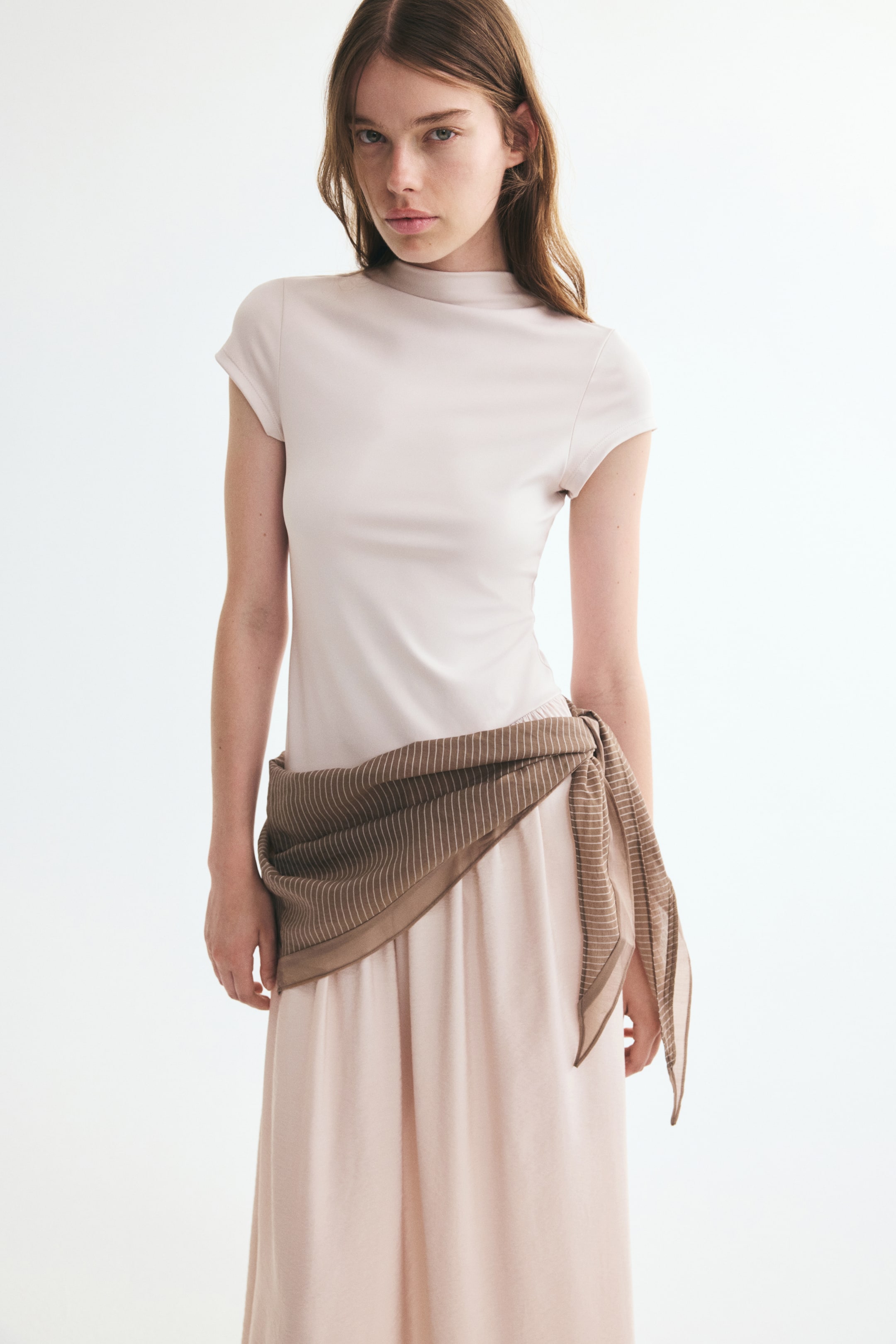 View larger image: The model is wearing a light beige maxi dress featuring a fitted bodice with a stand collar, cap sleeves, and a flared skirt. A brown striped scarf is tied around the hips over the dress’s asymmetrical waist seam.