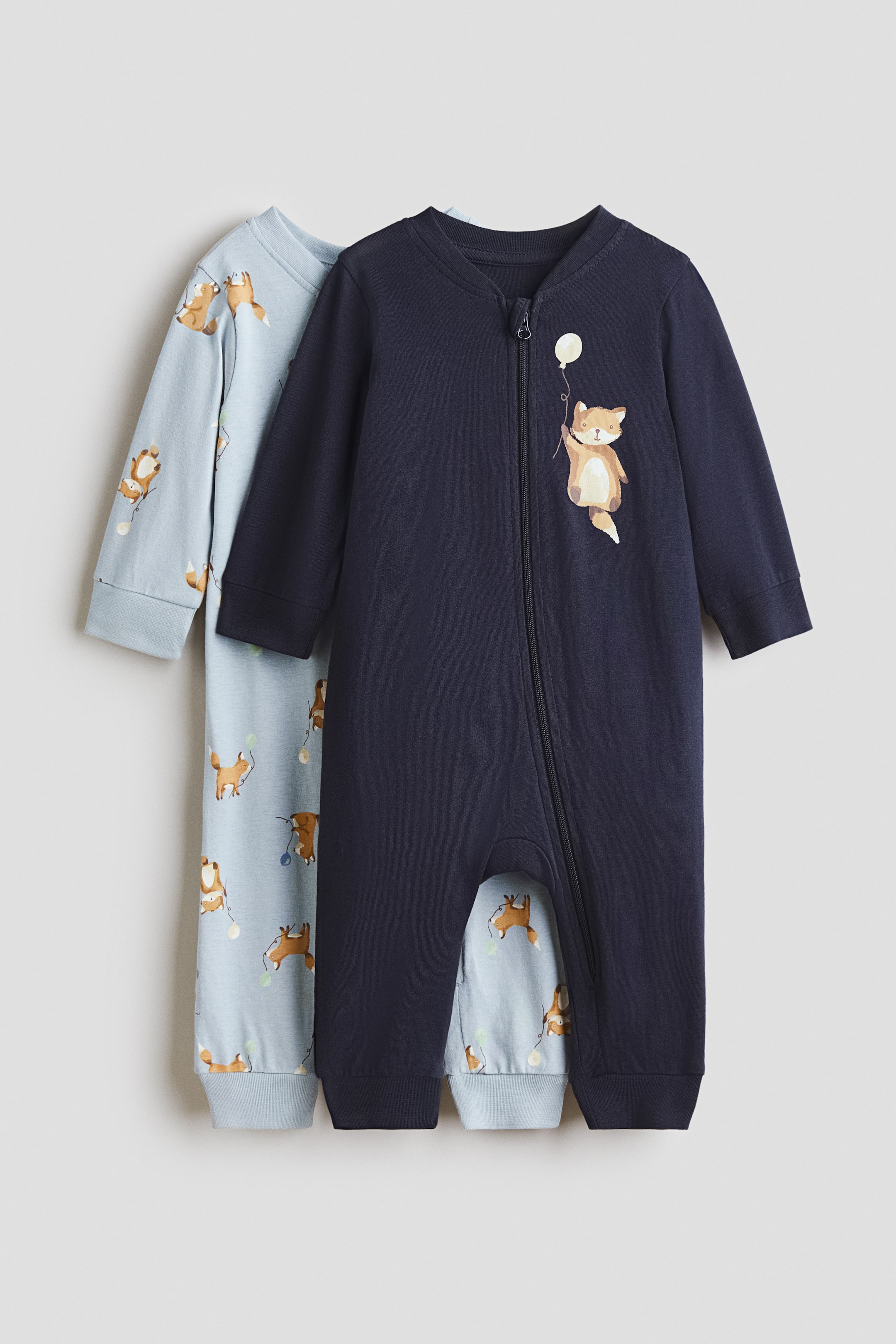 View larger image: Two baby sleepsuits are being presented, one slightly overlapping the other. The front sleepsuit is dark blue, featuring a central zip running to the crotch, a fox holding a white balloon on the chest, and simple cuffs on the sleeves and legs. The light blue sleepsuit behind it is patterned with an all-over print of foxes holding balloons and also has similar cuffs.