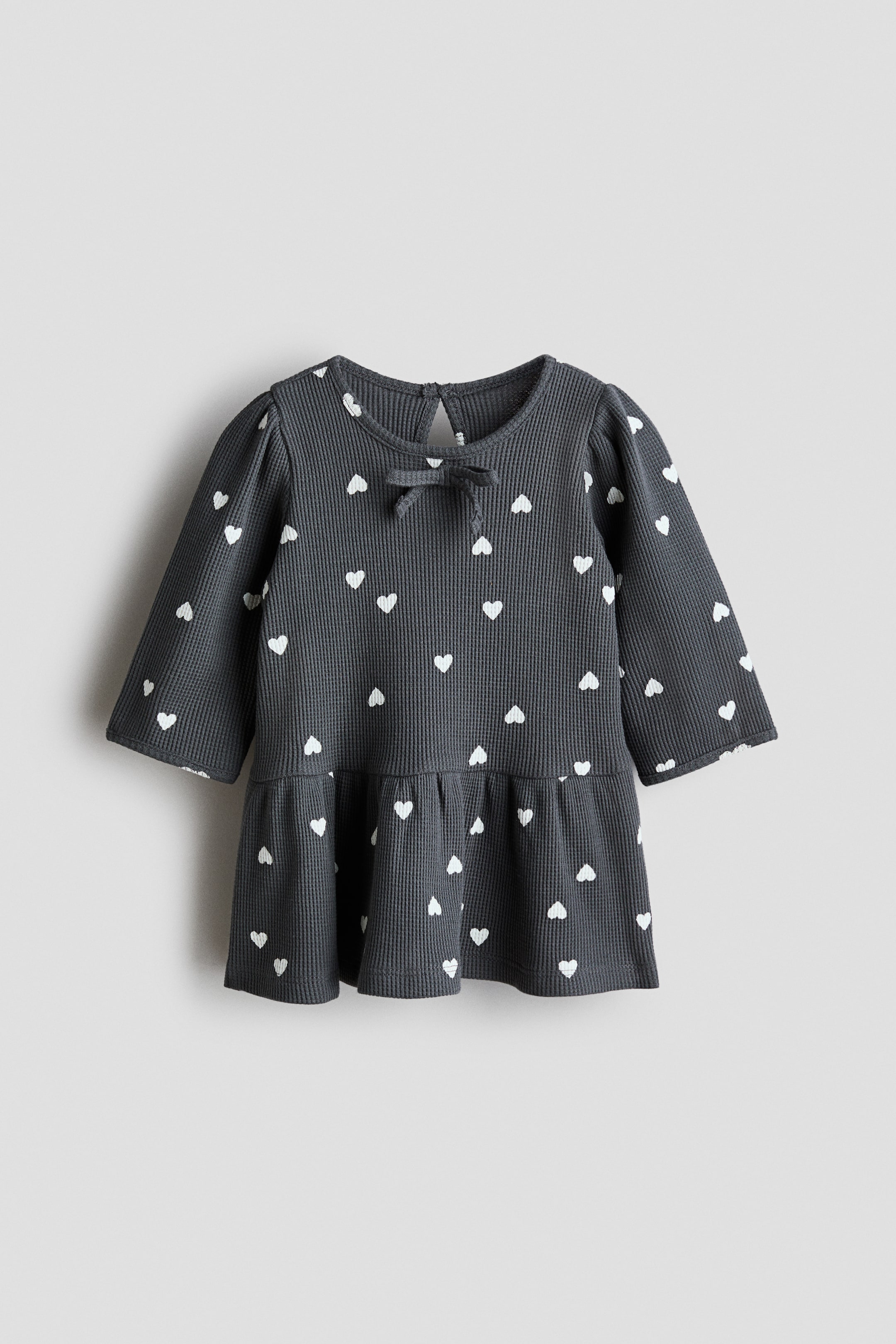 Agrandir l'image: Une robe pour bébé en côtes de couleur gris foncé présente un imprimé de cœur blanc et est montrée de face. Elle est conçue avec un col rond, un petit nœud décoratif à l'avant et une ouverture en goutte d'eau à l'arrière. Elle met en avant des manches longues avec des épaules légèrement froncées et un ourlet péplum à volants.