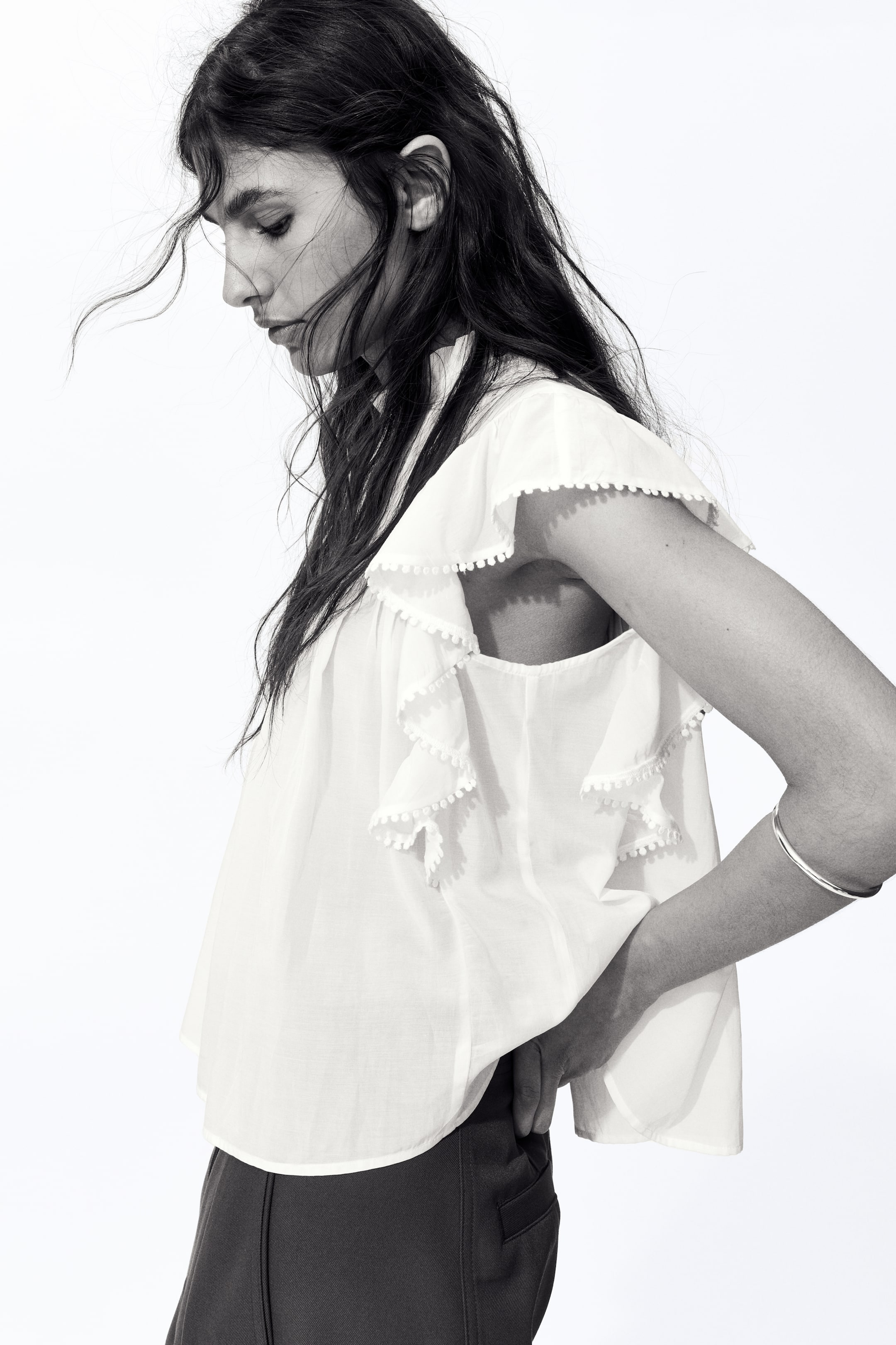 View larger image: A white top featuring tiered ruffled sleeves and a loose, flowing silhouette. Each ruffle is edged with small pom-poms, and the top boasts a curved hemline.