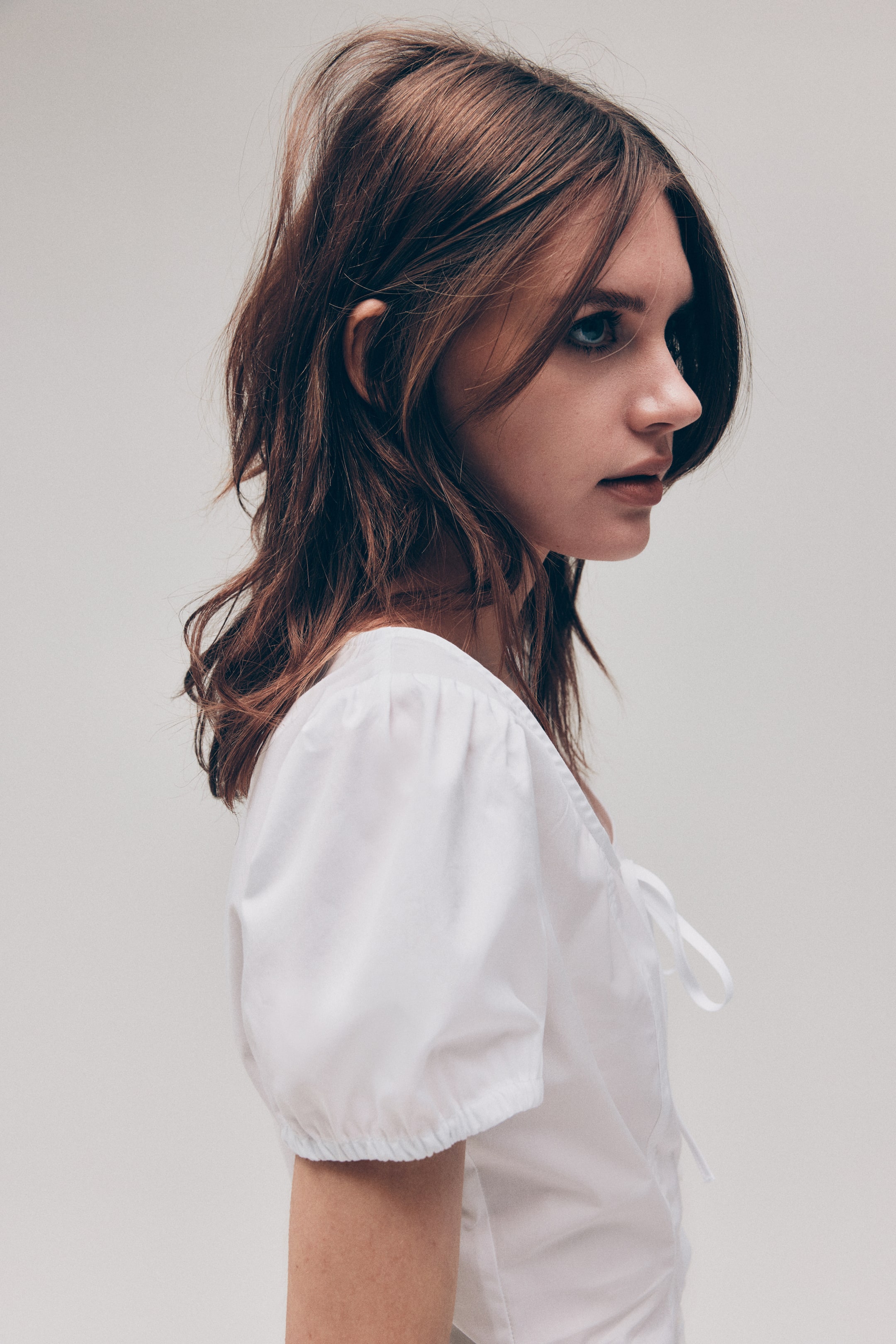 View larger image: The model is wearing a white cotton poplin blouse with a scoop neckline and narrow ties at the front, finished with short puff sleeves and elasticated cuffs.