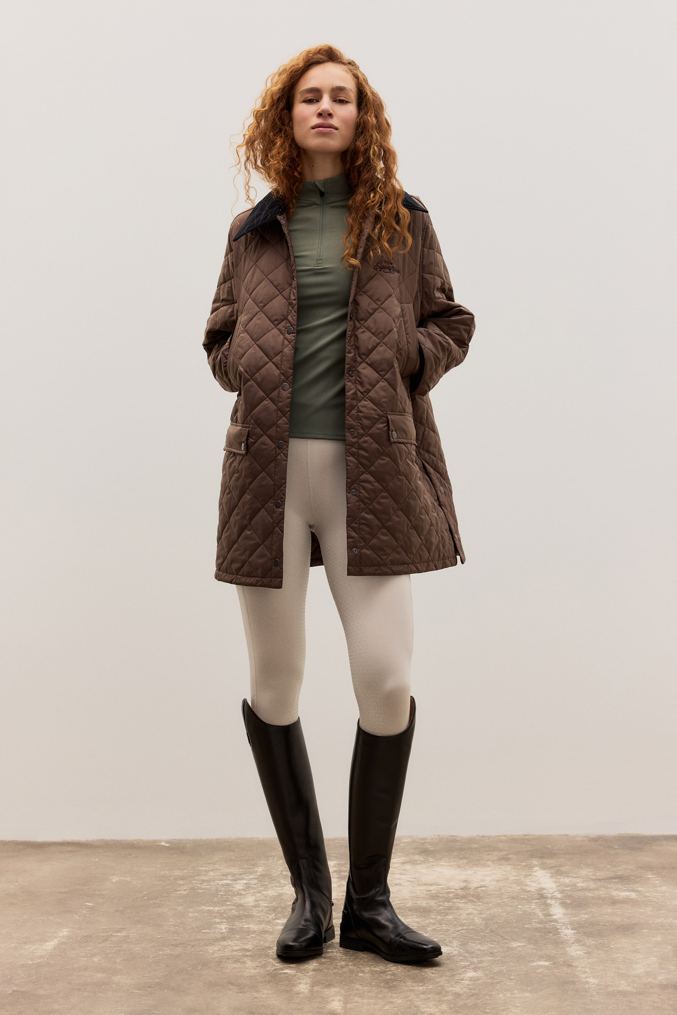 Grotere afbeelding bekijken: Een bruine doorgestikte jas met een ruitpatroon, een donkere contrasterende kraag, knoopsluitingen en klepzakken, wordt gedragen over een olijfgroene half-zip longsleeve top, licht beige rijbroek en zwarte hoge rijlaarzen, allemaal naar voren gericht gepresenteerd.