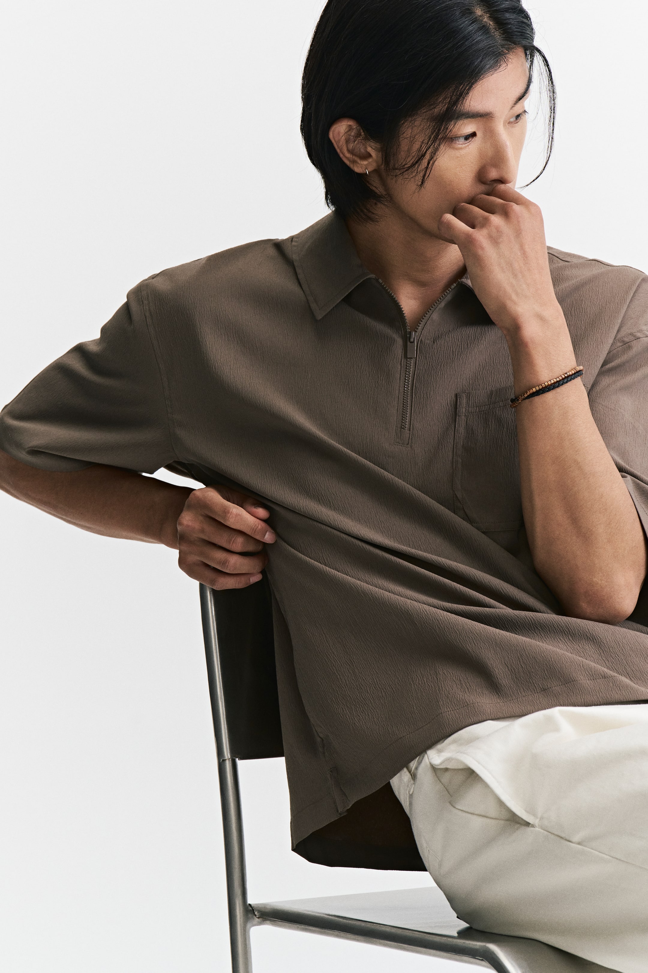 View larger image: The model is wearing a brown polo shirt in a crinkled weave, featuring a collar, half-zip closure, short sleeves, and a chest pocket. Styled with white trousers, the loose-fitting shirt is shown as the model sits on a metal chair.