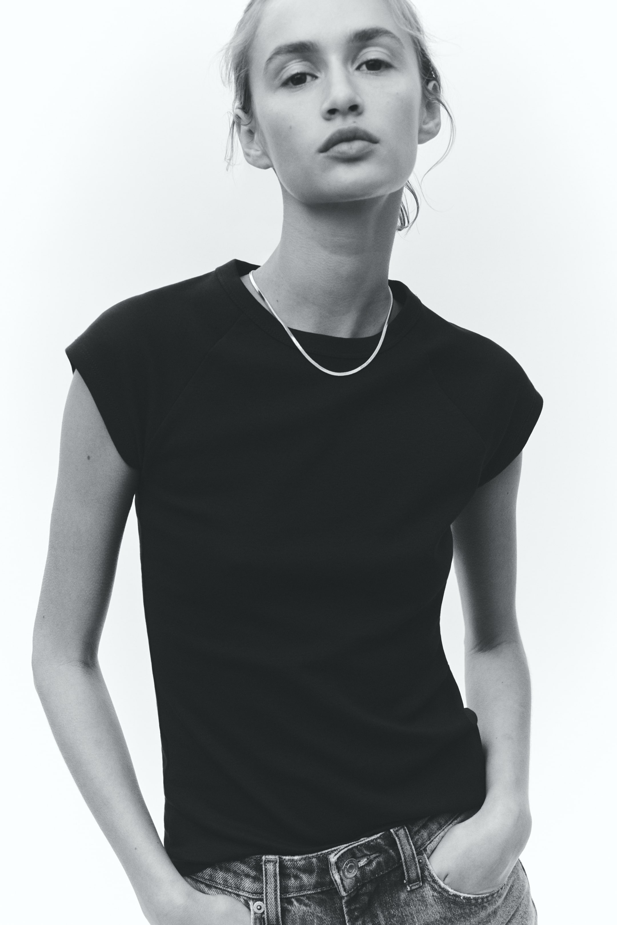 View larger image: The model is wearing a fitted t-shirt with a round, ribbed neckline and short raglan sleeves. A delicate chain necklace lies on top of the t-shirt, while the model's hands are casually placed in her jean pockets, subtly altering the shirt's drape.